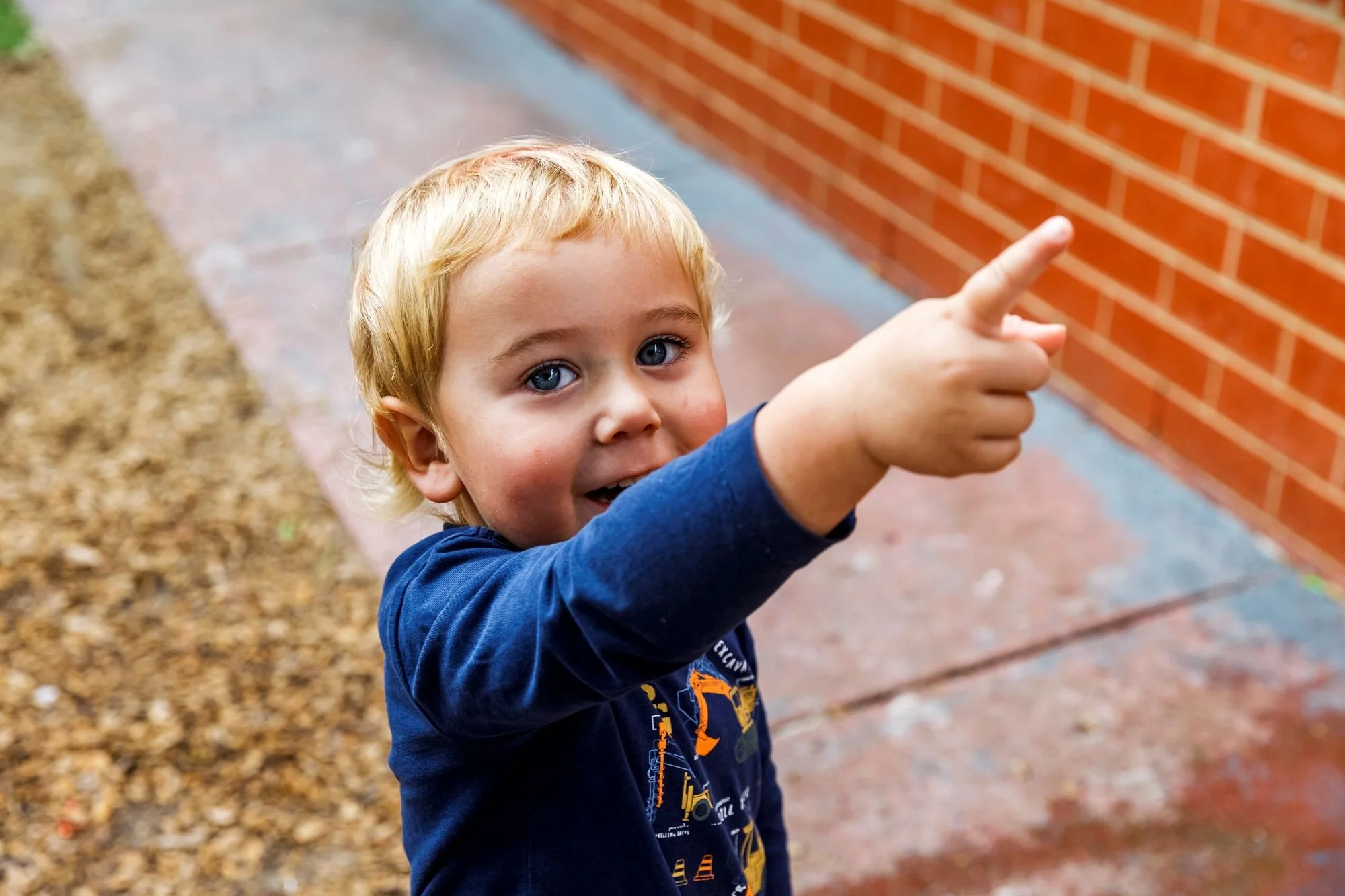 A young boy with blonde hair and blue eyes pointing to his right while smiling, outdoors near a brick wall.