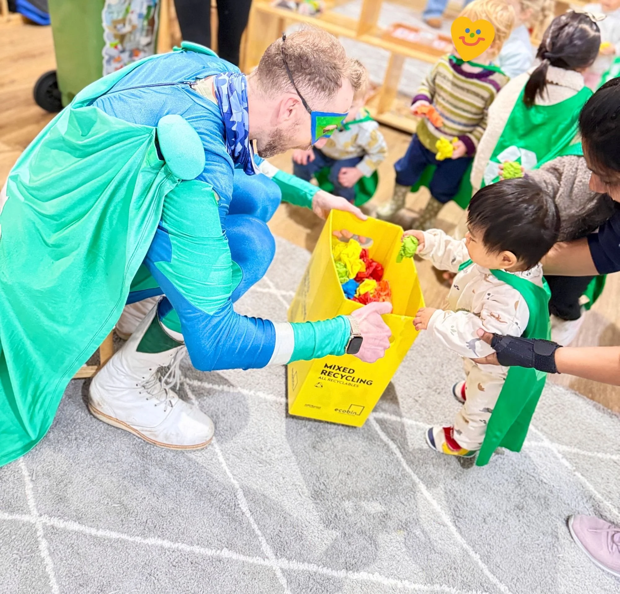 Children at a recycling station participating in an activity, supervised by an adult wearing a blue and green costume, with a yellow recycling bin labeled "Mixed Recycling".