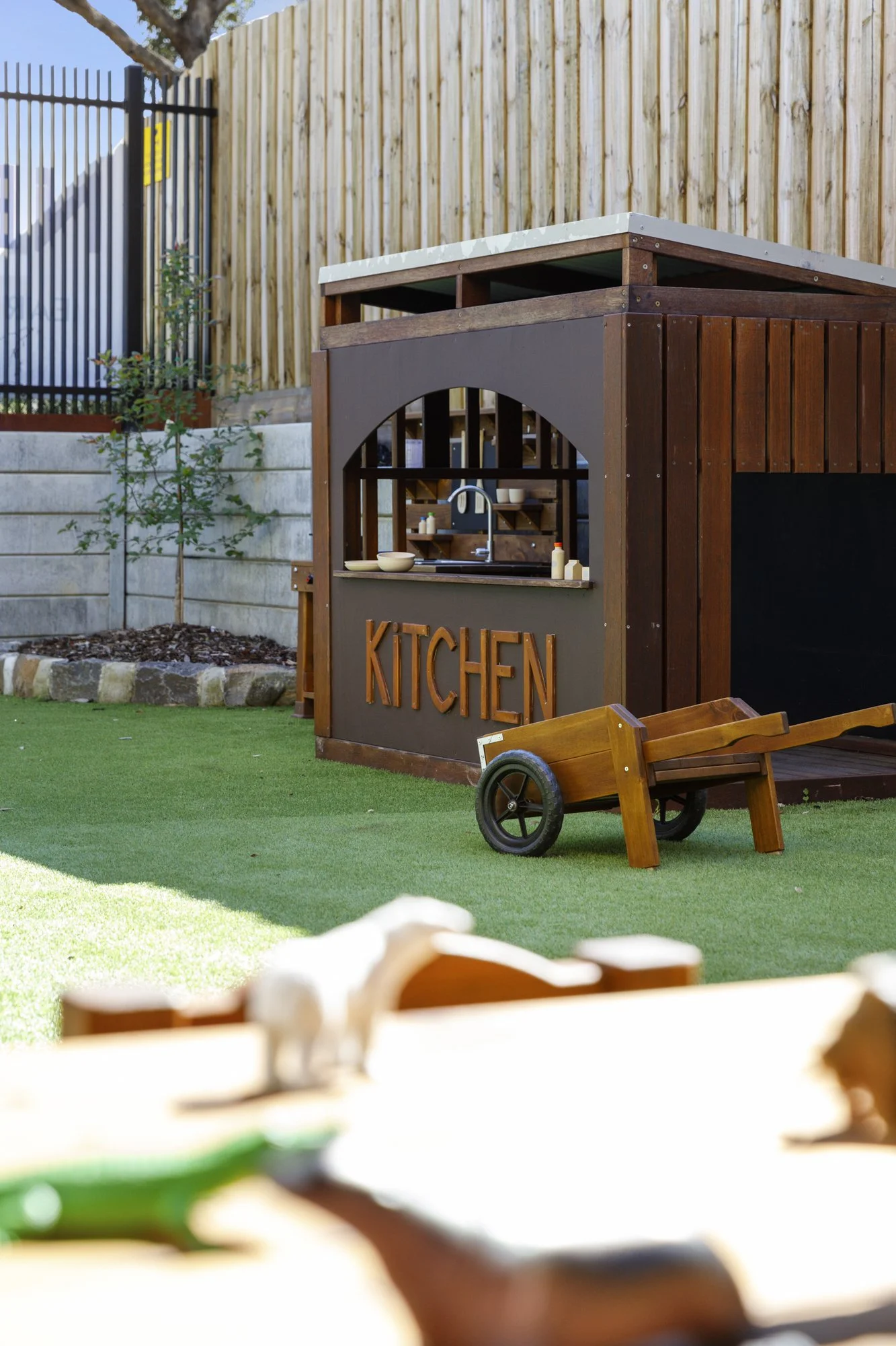 Backyard with a wooden playhouse labeled 'KITCHEN', a small wooden wheelbarrow, and a lush green lawn. There is a wooden fence, some plants, and a tree in the background.