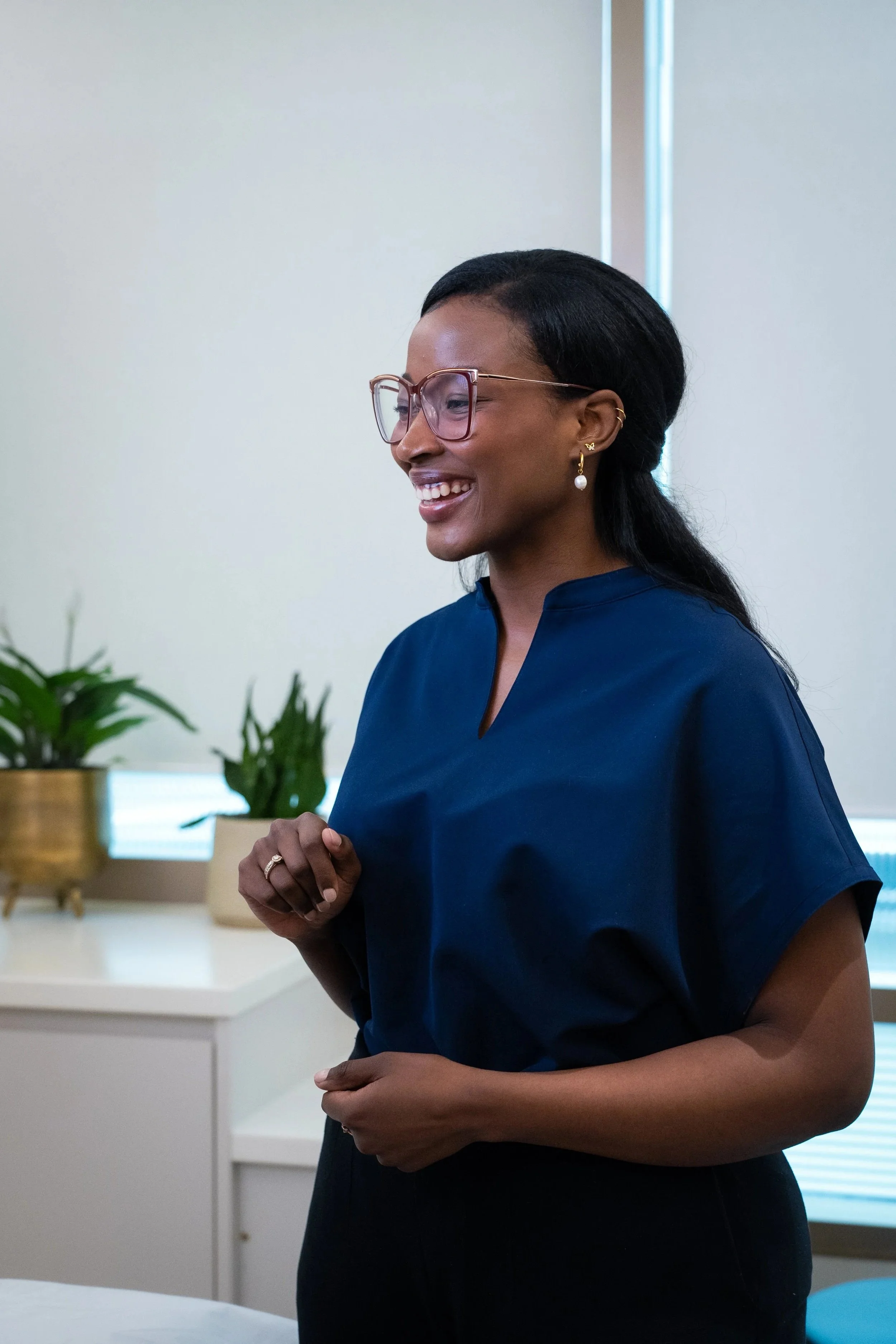 A smiling woman with glasses and earrings, wearing a blue scrub top, standing indoors with plants and a white cabinet in the background.