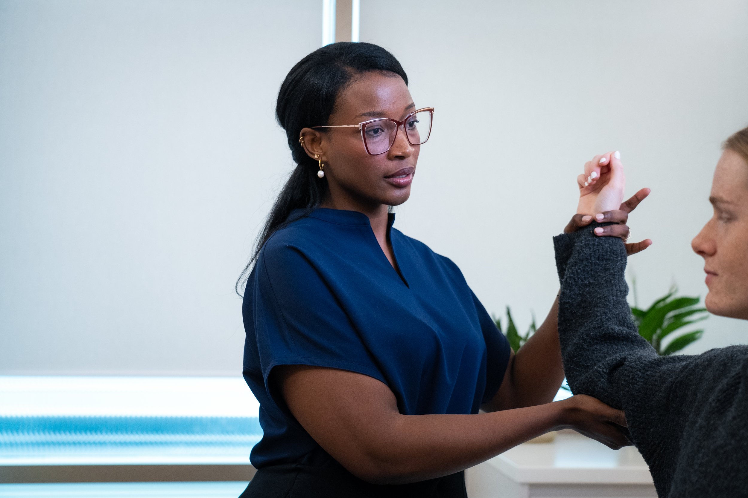 A woman with glasses and earrings in a blue top appears to be performing a physical examination or therapy on a person's arm, which is raised and bent at the elbow.