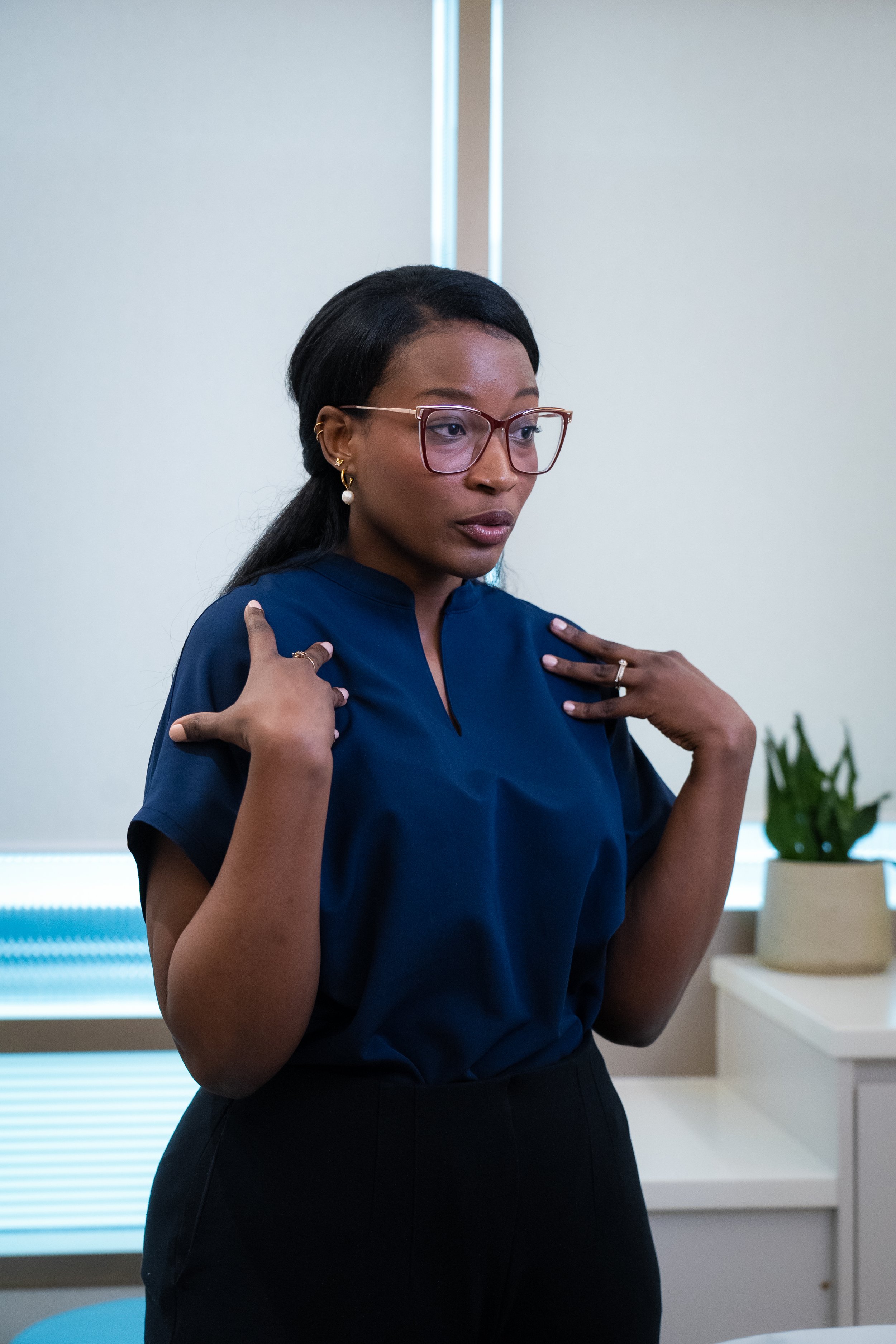 A woman with dark hair and glasses, wearing a blue blouse and black pants, standing in a treatment room, adjusting her shoulders with her hands.