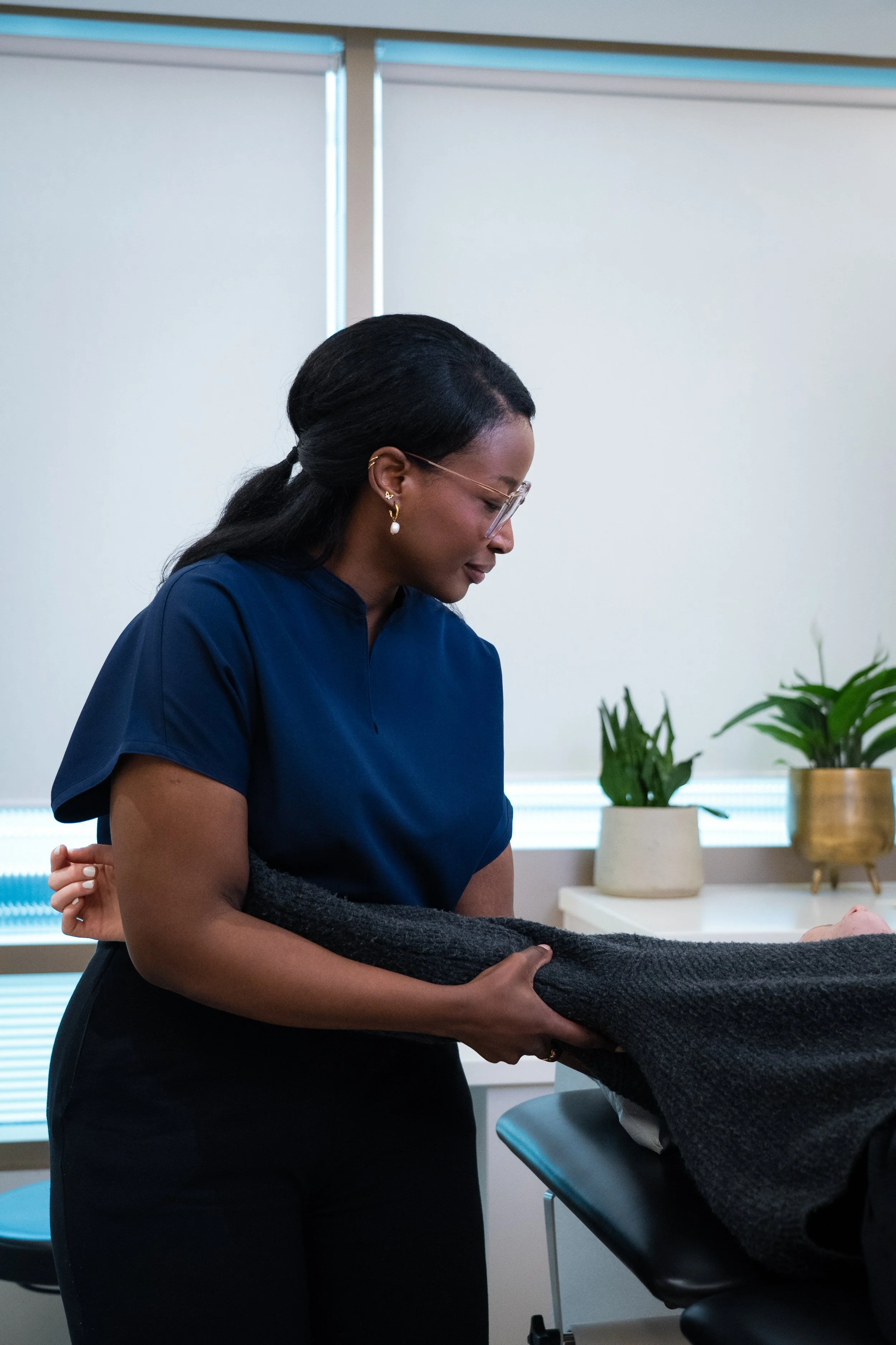 A woman dressed in navy scrubs is handing a black blanket to a patient in a medical setting with potted plants and large windows in the background.