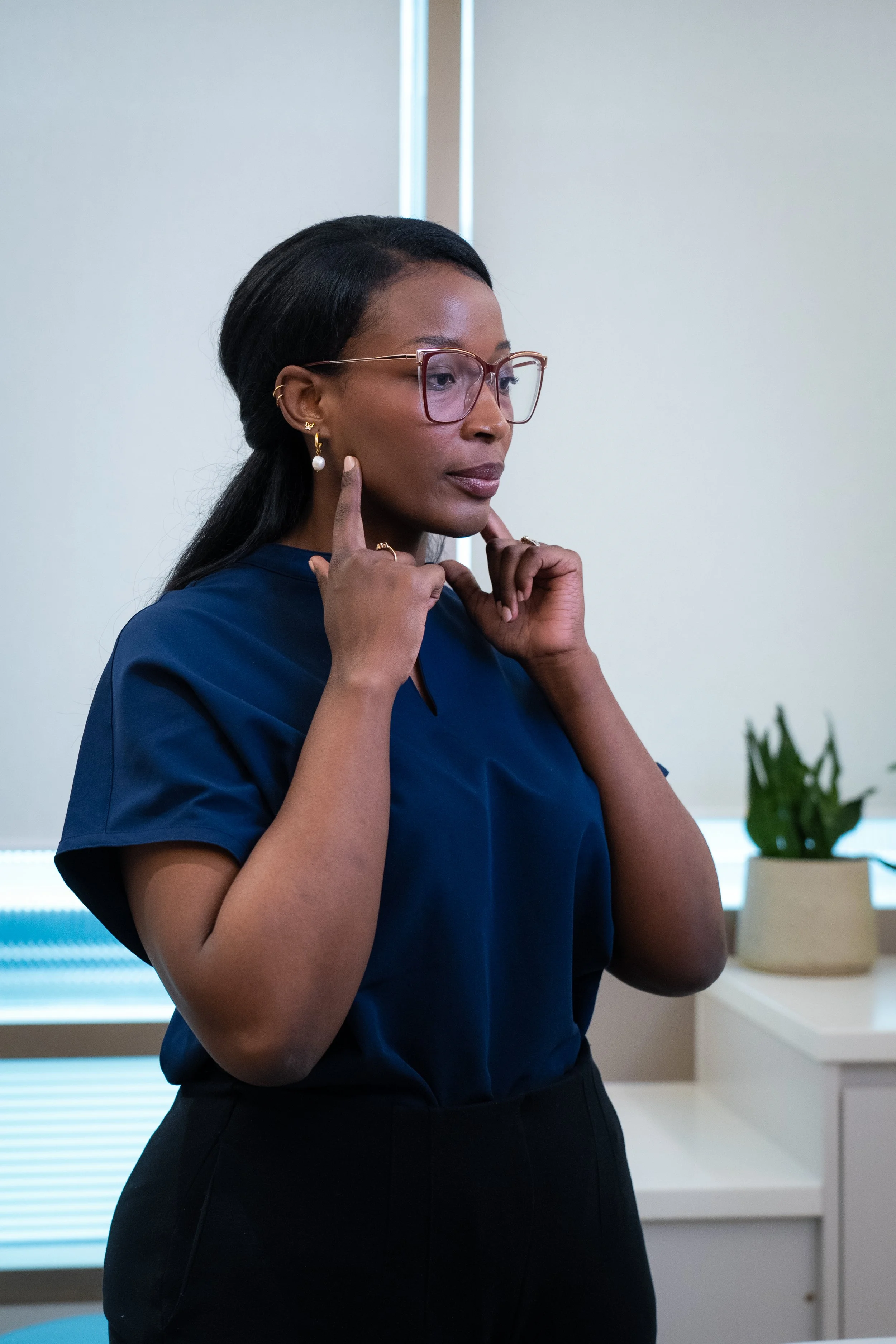 A woman in glasses and professional attire touching her face with both hands in an office setting.