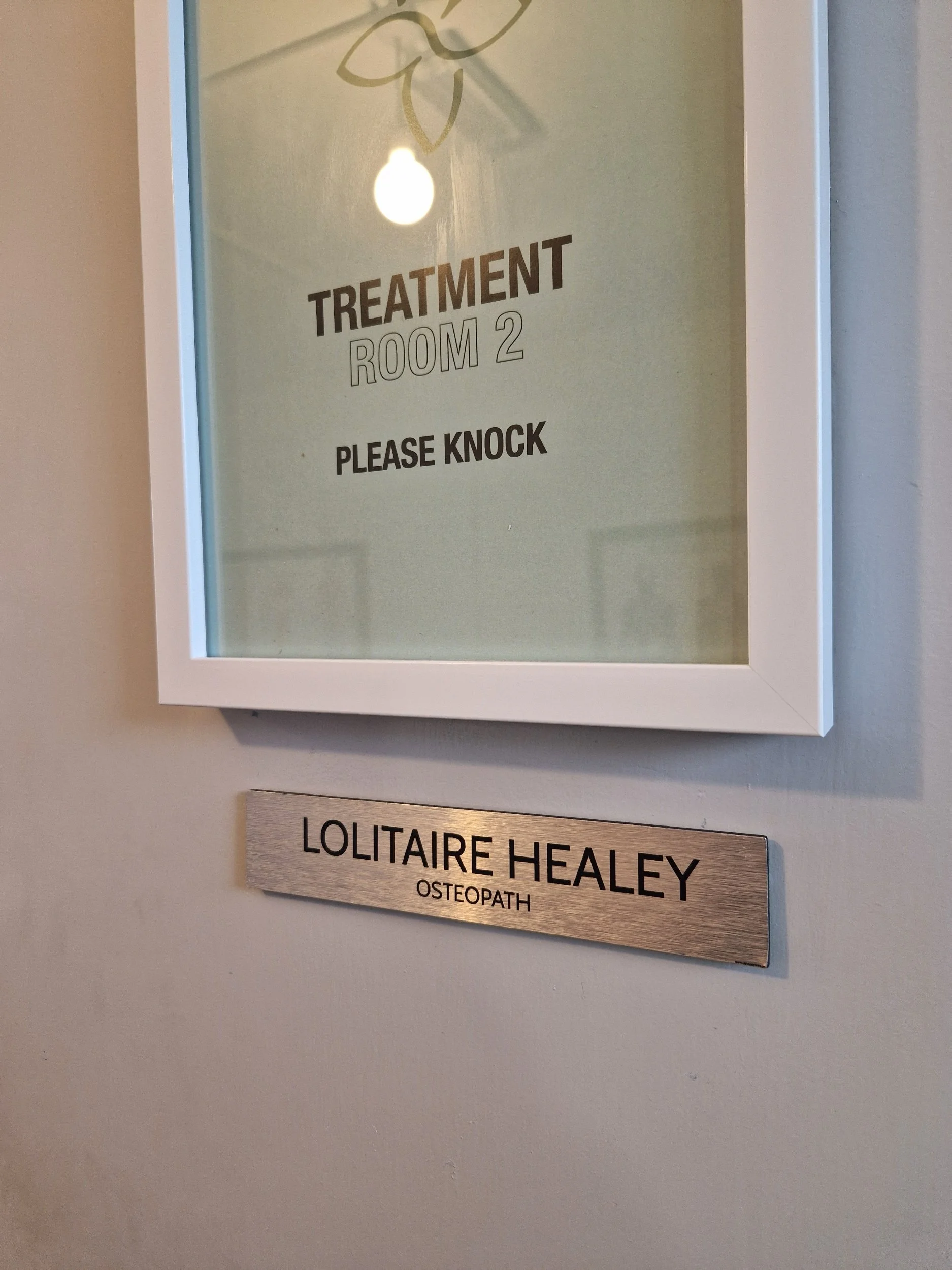 A door sign indicating a treatment room labeled 'Treatment Room 2 Please Knock' with a nameplate below that reads 'Lolitaire Healey Osteopath'.