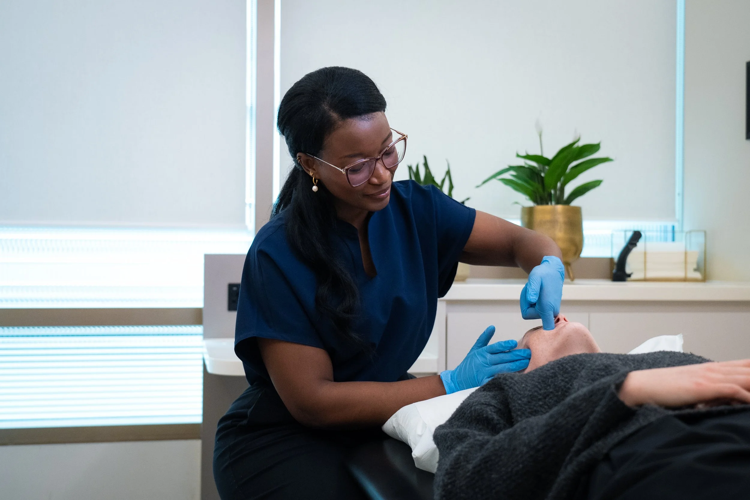 A woman in blue scrubs and glasses performs jaw treatment on a patient lying on a treatment bed in a medical office.