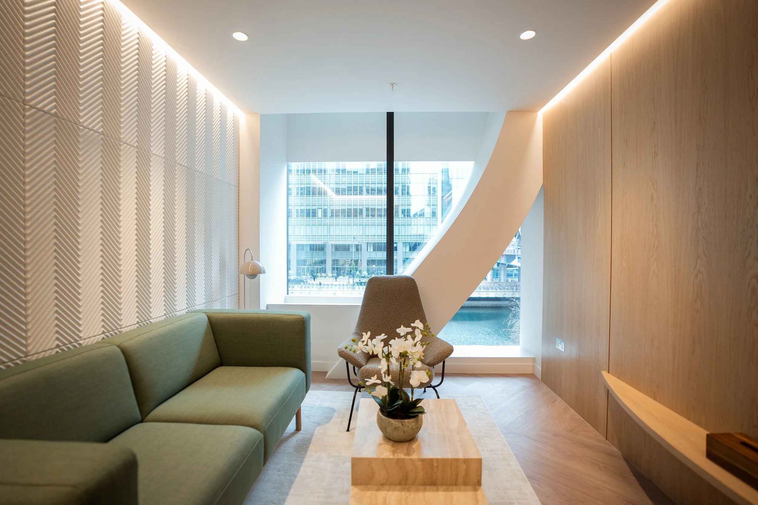 Modern coaching room with a green sofa, a single beige armchair, a small wooden coffee table with a potted orchid, a large window with cityscape view, and warm wooden wall accents.