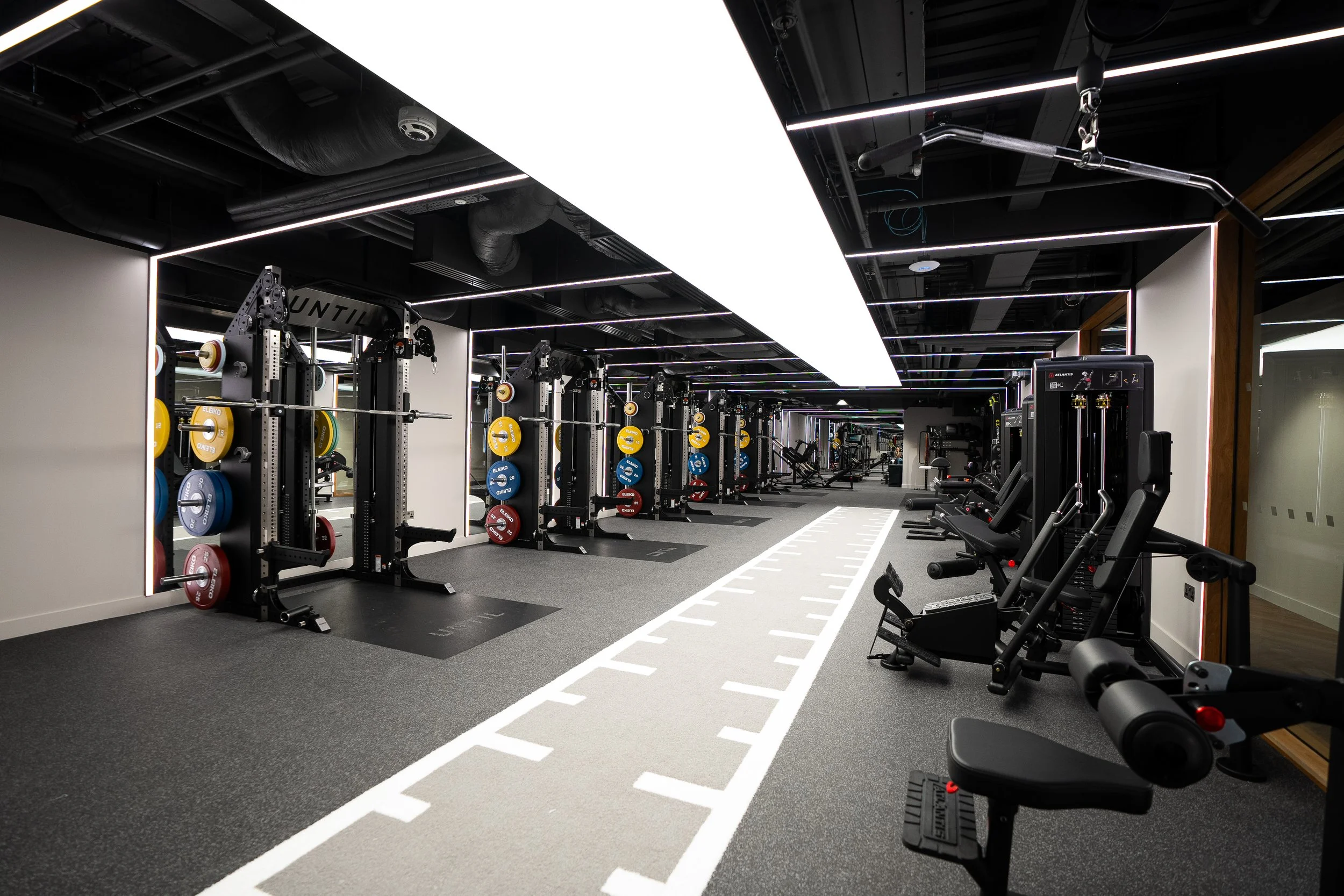 Modern gym with various workout equipment including squat racks, weight plates, and exercise machines, illuminated by bright ceiling lights.