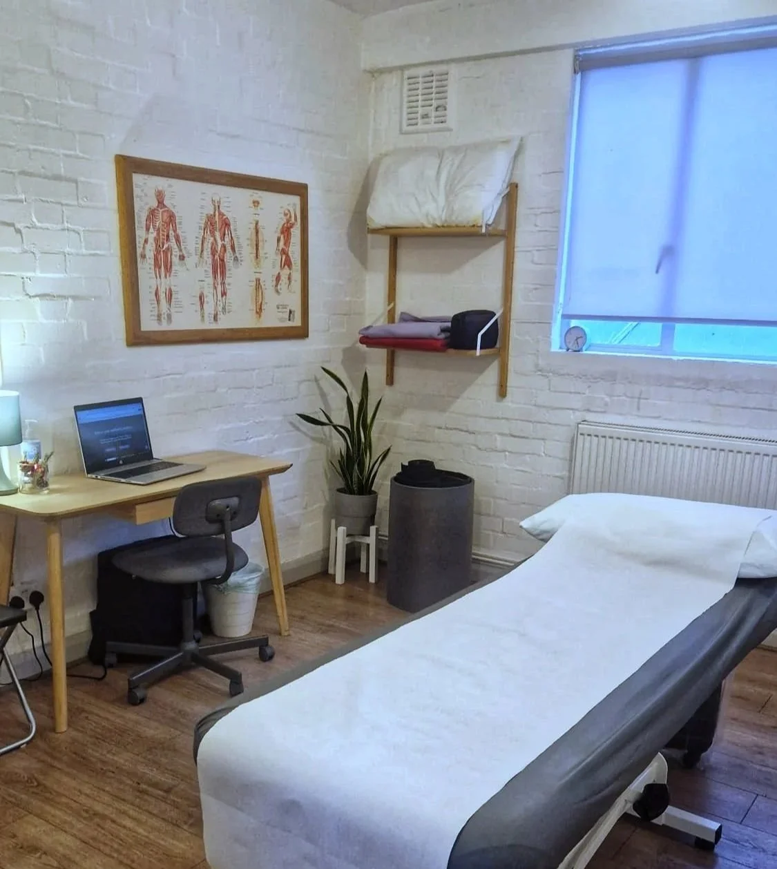 Medical examination room with a bed, a computer on a wooden desk, a black rolling chair, a chart of human muscles on the wall, a plant, and shelves with folded linens, a clock, and a window with a blue shade.