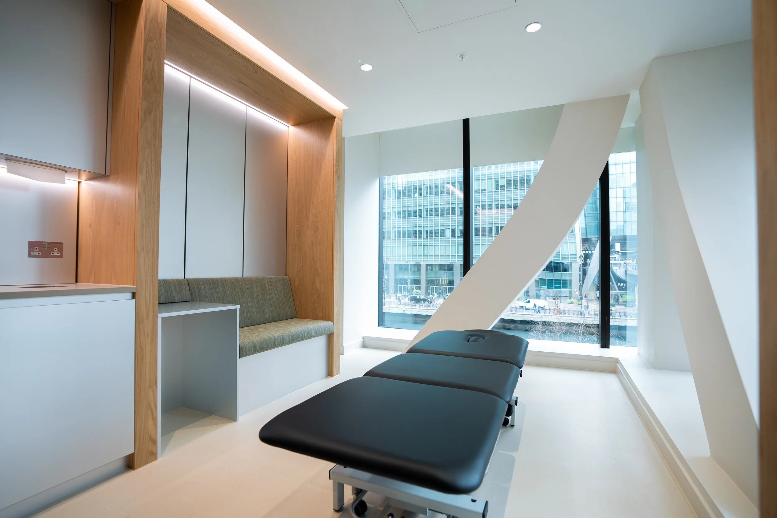 Modern medical examination room with reclining examination table, built-in seating, large windows, cityscape view, and minimalist decor.