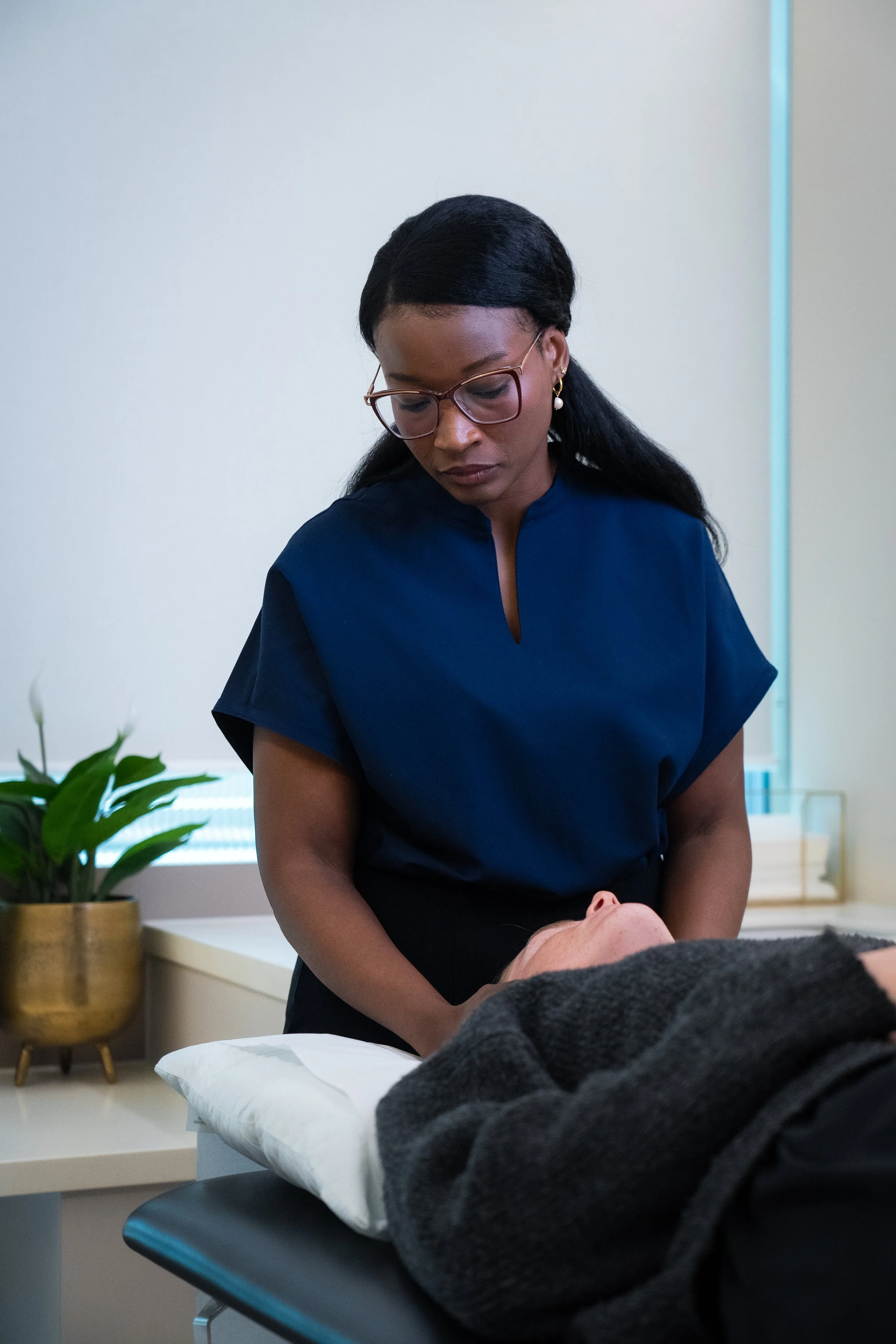 A woman with glasses and earrings provides Osteopathic care to a person lying face up on a treatment table in a clinic room, with a potted plant in the background.