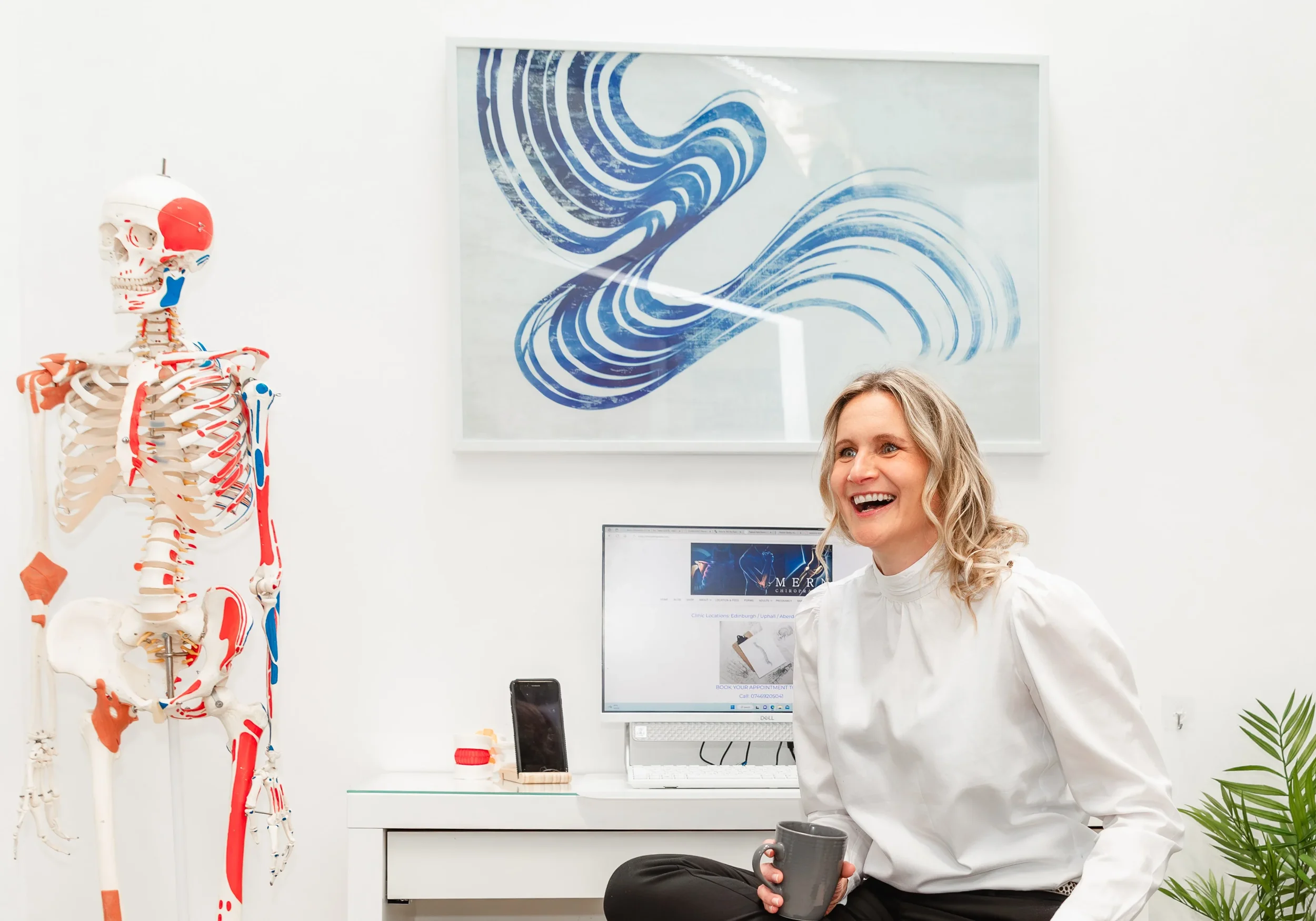A woman with blonde curly hair is smiling and holding a gray mug in her right hand, sitting in an office with a human skeleton model to her left and a computer monitor in front of her. On the wall behind her, there's an abstract blue wave artwork, and to her left, there's a plant.
