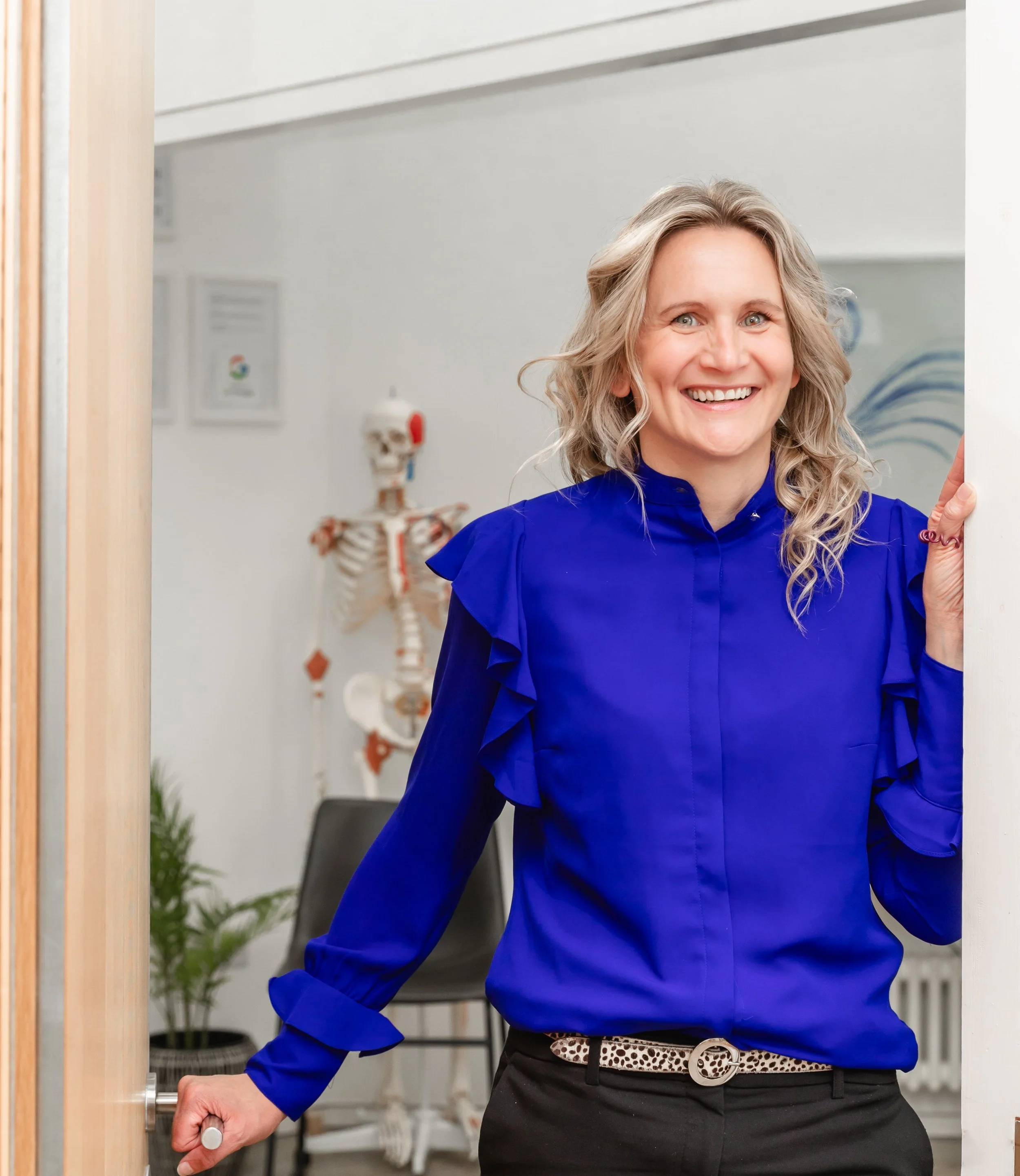 A woman opening a door, smiling, wearing a royal blue blouse, with a leopard print belt and black pants, in a room with medical posters and a skeleton model in the background.