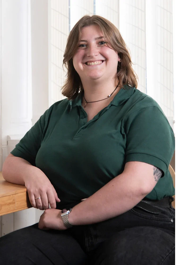 A smiling woman with shoulder-length brown hair, wearing a dark green polo shirt and jewelry, sitting with her arm on a wooden table in front of large windows.