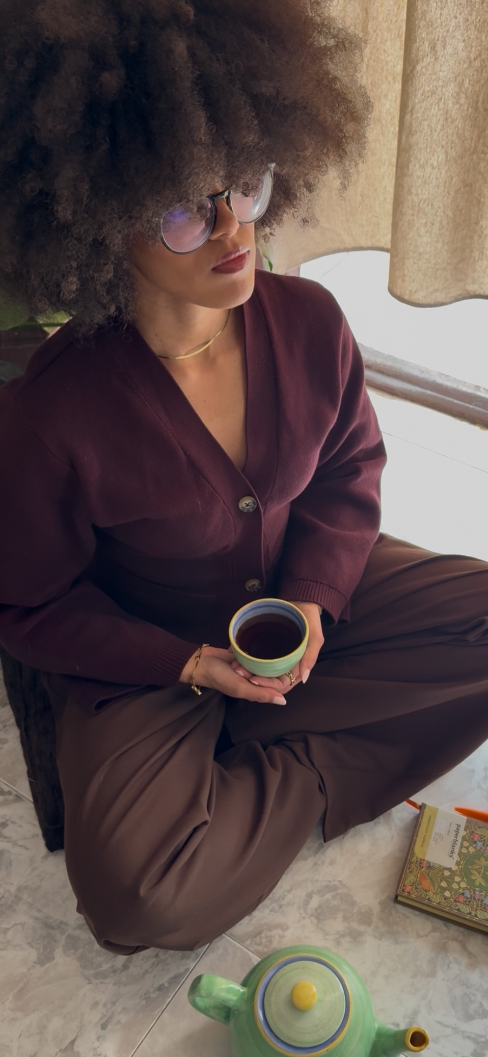 Eine Frau mit großer Afro-Frisur, Sonnenbrille, dunkelrotem Blazer und braunen Hosen sitzt auf dem Boden, hält eine Tasse Kaffee und trinkt. Es ist eine gemütliche Szene am Fenster mit einem Buch und einem grünen Teekessel auf dem Boden.