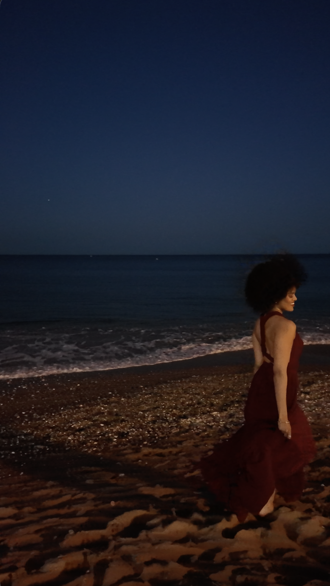 Eine Frau in einem roten Kleid läuft barfuß am Strand entlang, während die Nacht hereinbricht, mit dunklem Himmel und Meereswellen im Hintergrund.