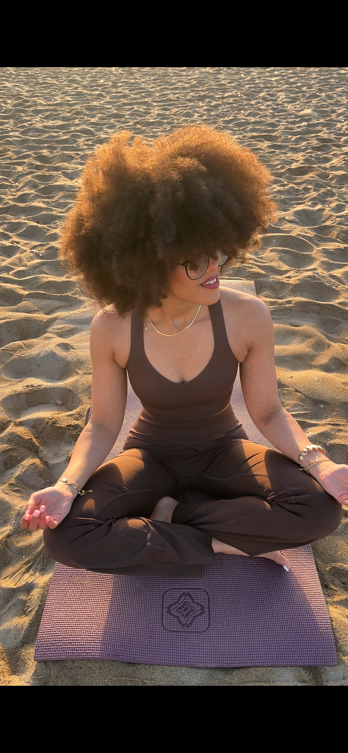 Eine Frau sitzt in einer Yoga-Position auf einem Yoga-Matte am Strand im Sonnenuntergang, trägt eine schwarze Sportbekleidung und hasst lockiges, volles Haar.