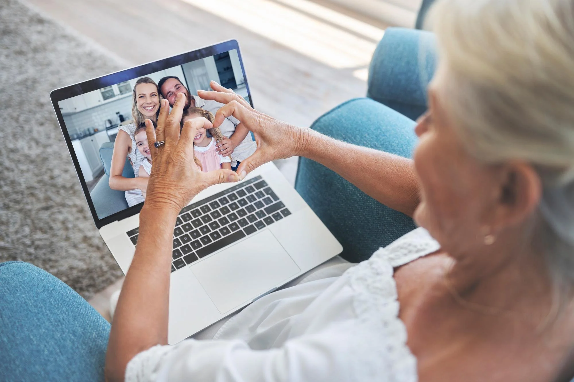 Older woman with blonde hair making a heart shape with her hands in front of a video call on a laptop, showing a happy family of four.