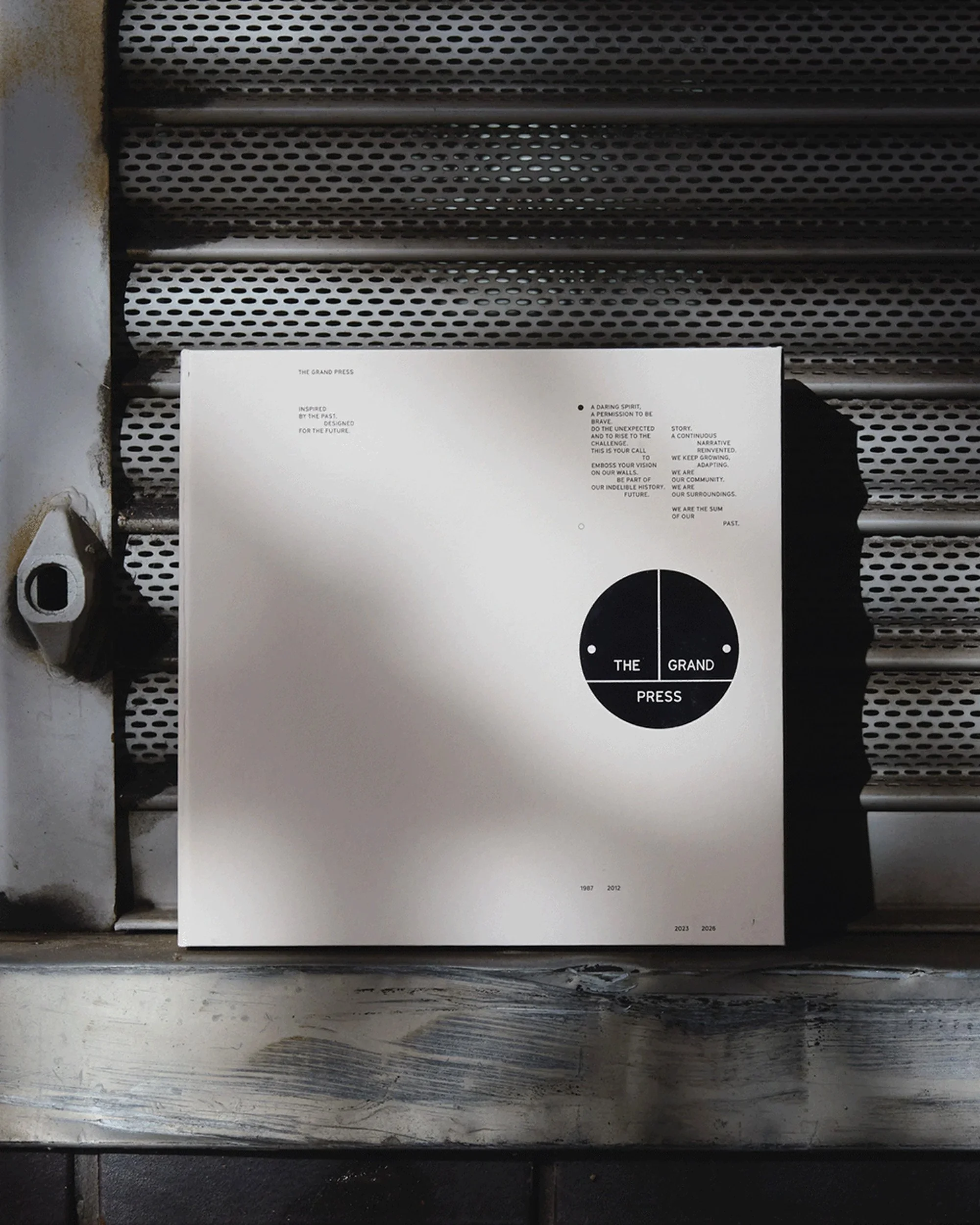 The Grand Press Guidelines. A white poster titled 'The Grand Press' leaning against a metal vent in an industrial setting. The poster features black text and a black circle divided into four quadrants with 'The,' 'Grand,' and 'Press'.