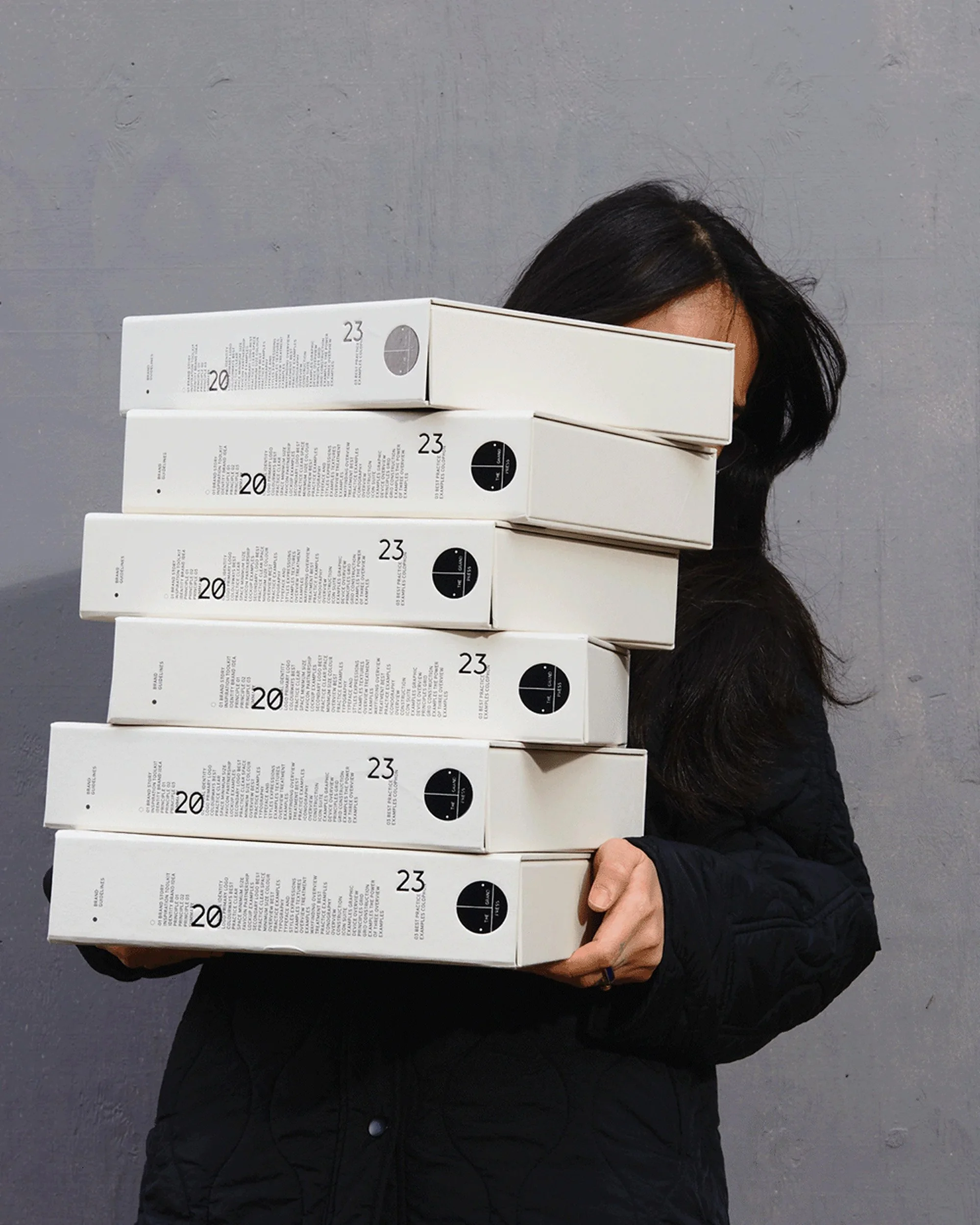 The Grand Press Guidelines. Person holding multiple stacked pizza boxes in front of a grey wall.