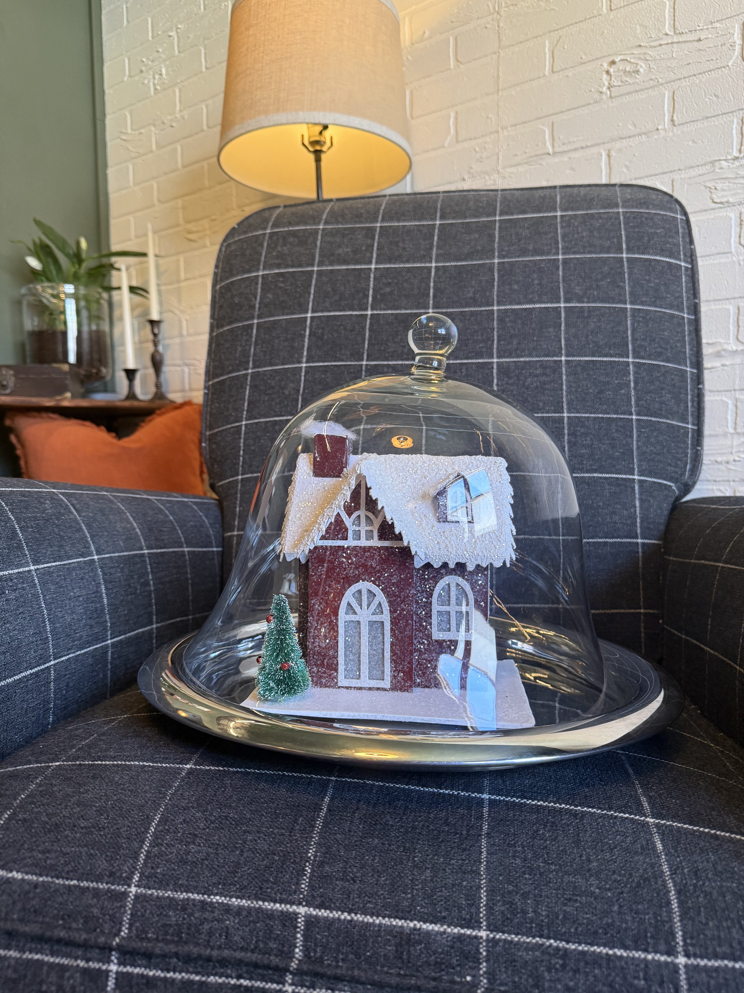 A small decorative house on a silver tray inside a glass dome, with a miniature Christmas tree next to it, on a dark checkered chair in a cozy living room.