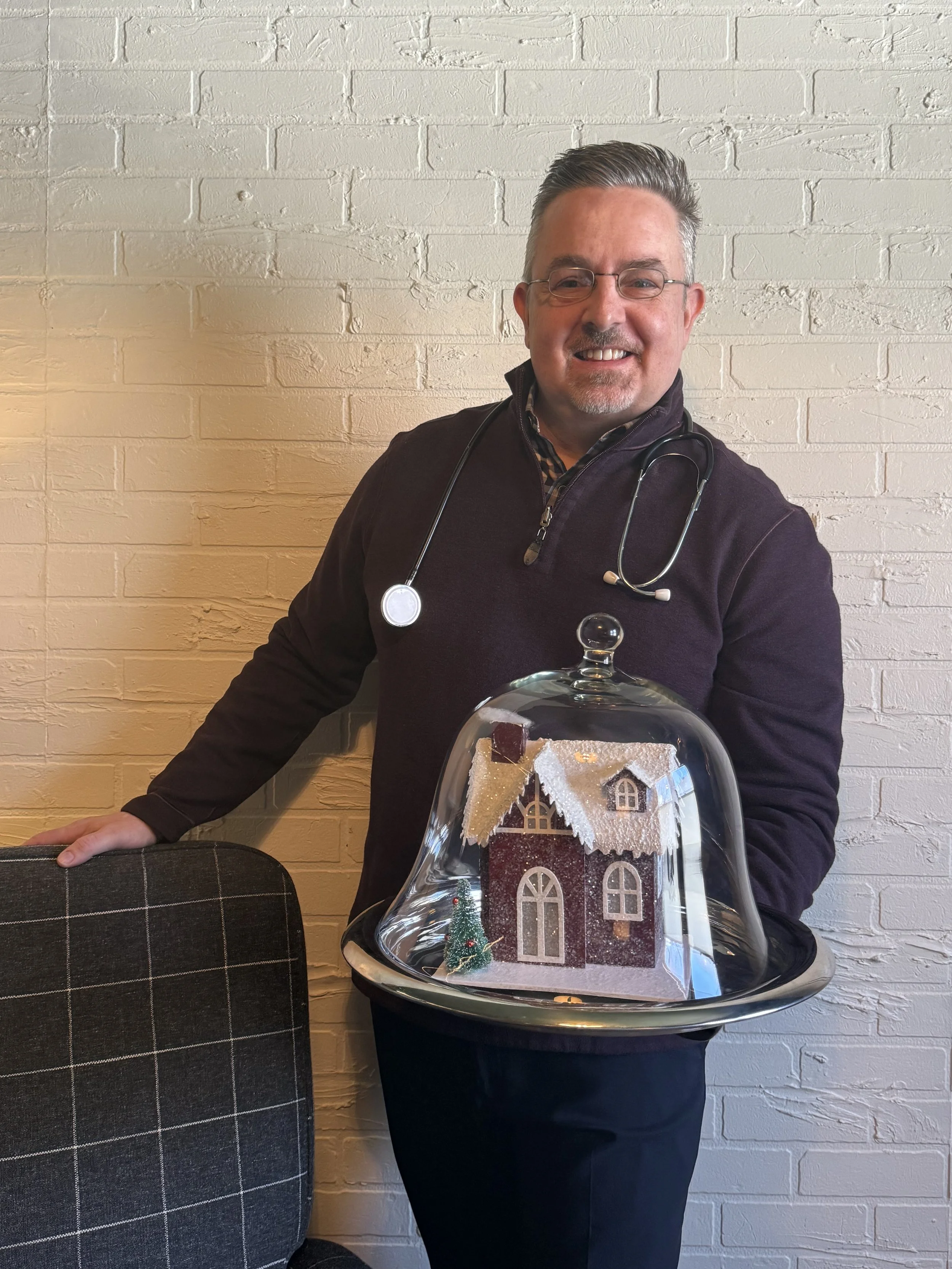 A man smiling, wearing glasses, a dark sweater, and a stethoscope around his neck, standing next to a chair against a white brick wall. He is holding a tray with a miniature snow-covered house cake under a glass dome.