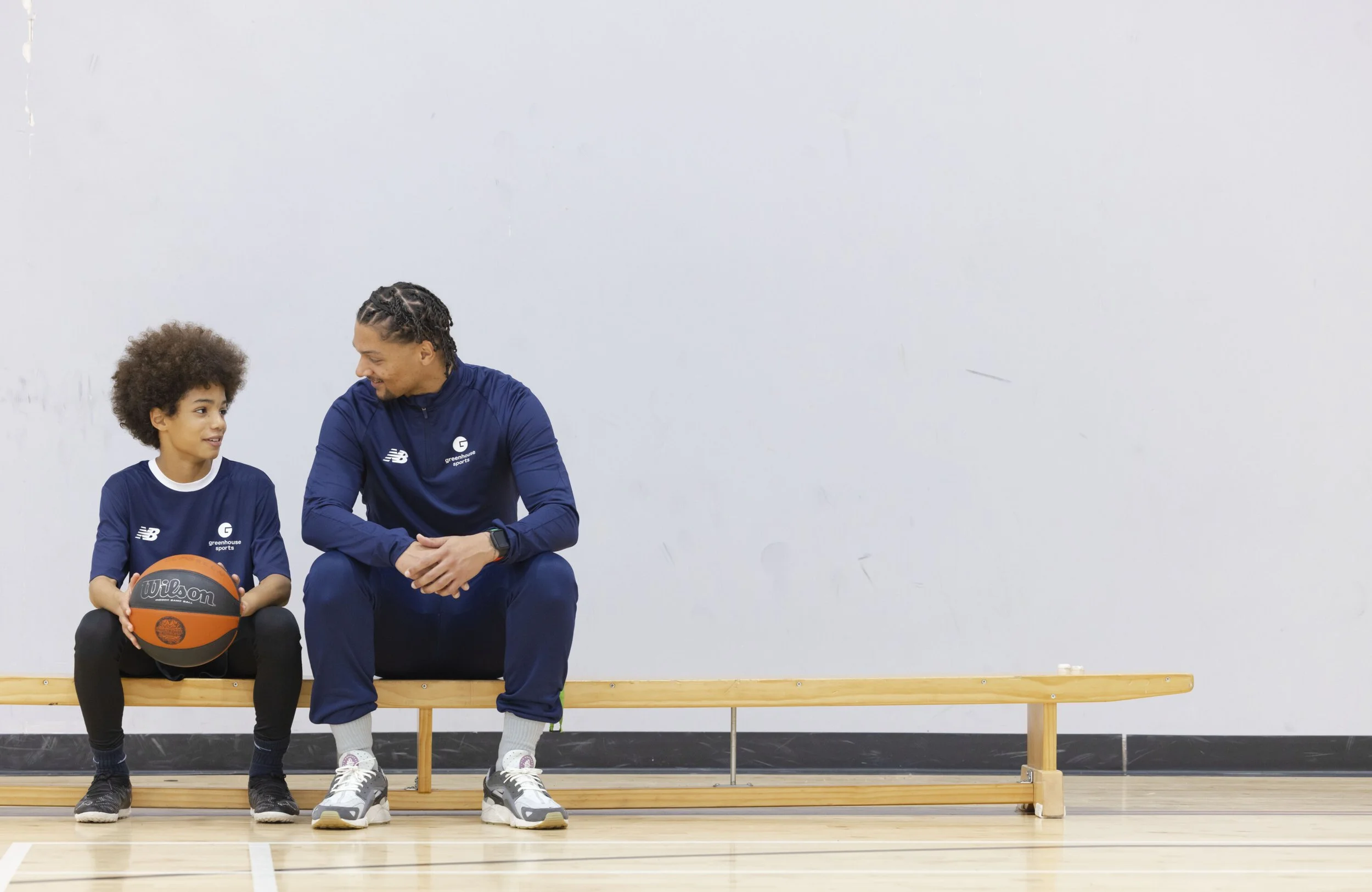 A young girl with curly hair holding a basketball, sitting on a bench next to a man with cornrows, both wearing matching blue sports outfits, indoors against a blank white wall.