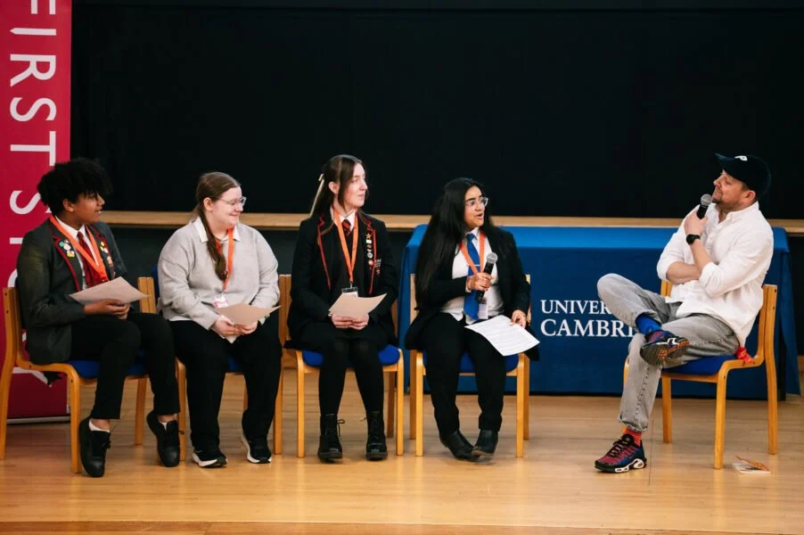 Five young women and one man sitting on stage in discussion, some holding microphones and notes, at the University of Cambridge, with a blackboard and university banner in the background.