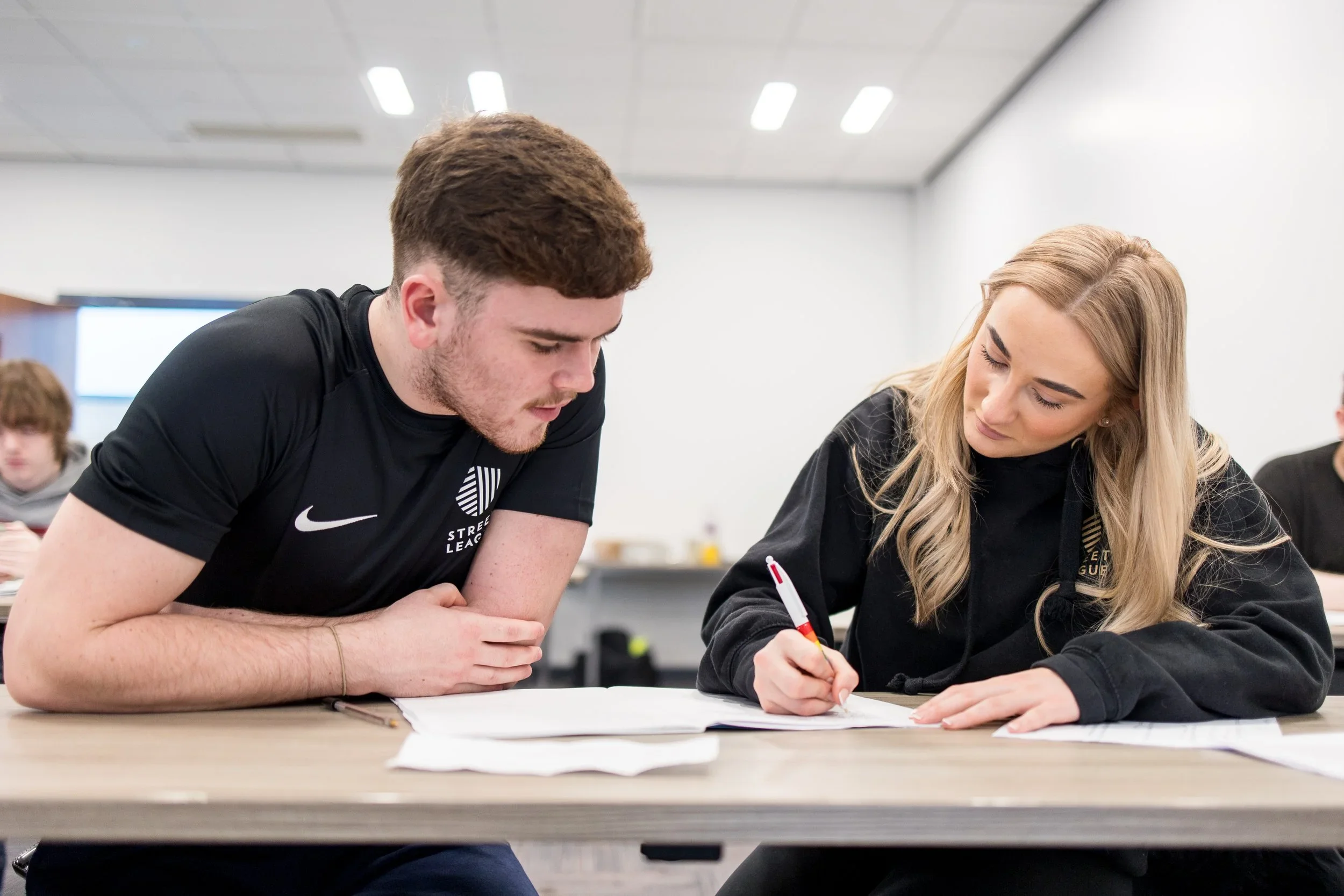 Two young adults, a man with short light brown hair and a woman with long blonde hair, are sitting at a table working together on a paper or worksheet, in a brightly lit room.