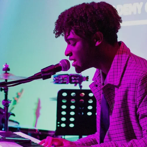 A young man with curly hair singing into a microphone while playing the keyboard on stage, with colorful lighting and a music stand in the background.