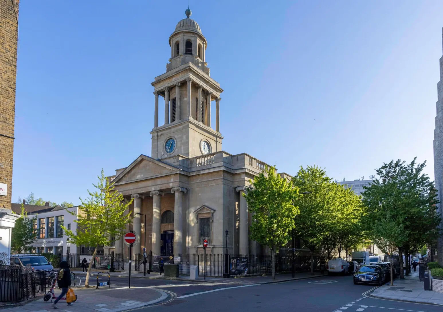 A historic church with a tall clock tower, columns, and a triangular pediment, located on a city street lined with green trees and parked cars, under a clear blue sky.