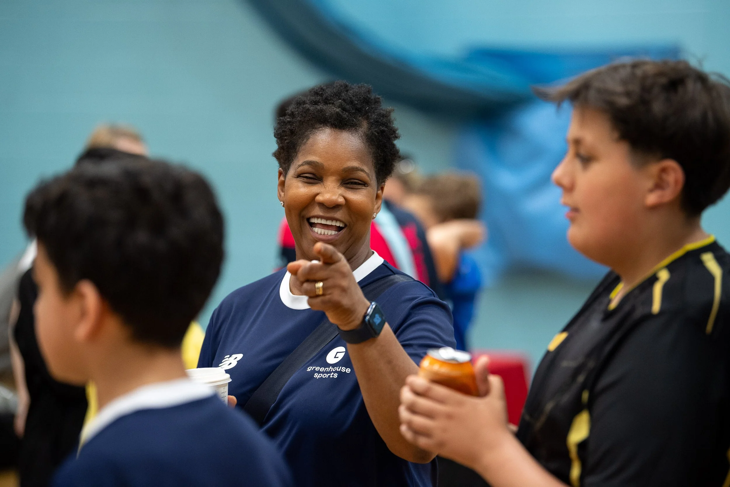 A woman with short curly hair smiling and pointing at a person in front of her, surrounded by others in a social setting.