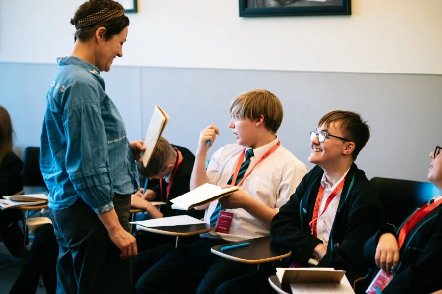 Teacher talking with students in a classroom, students are smiling and engaged in conversation.