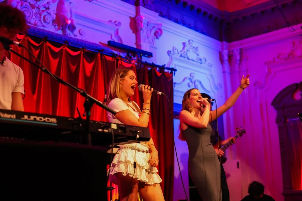 Two women singing into microphones on stage with a keyboard and guitarist, red curtains and ornate decorations in the background.
