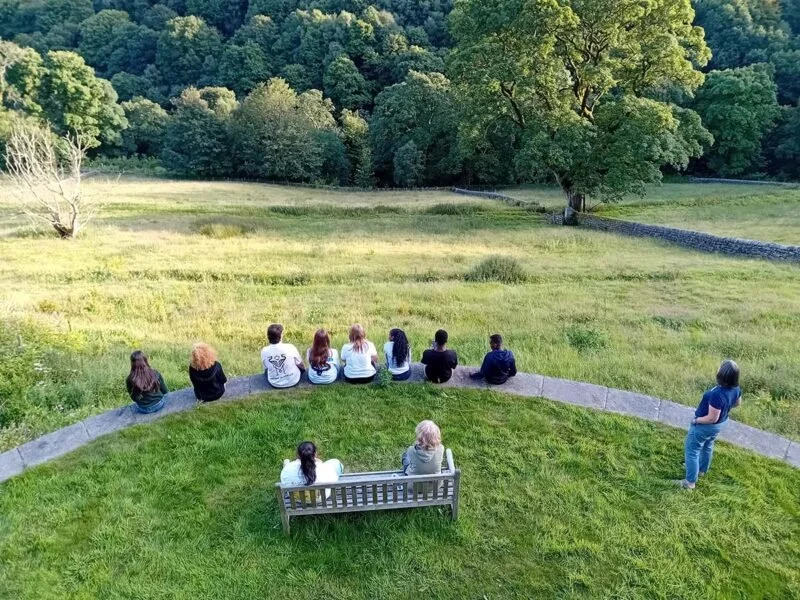 Ten people sitting on a curved bench and sitting on the grass, looking towards a grassy field with trees in the distance, with one person standing to the side.