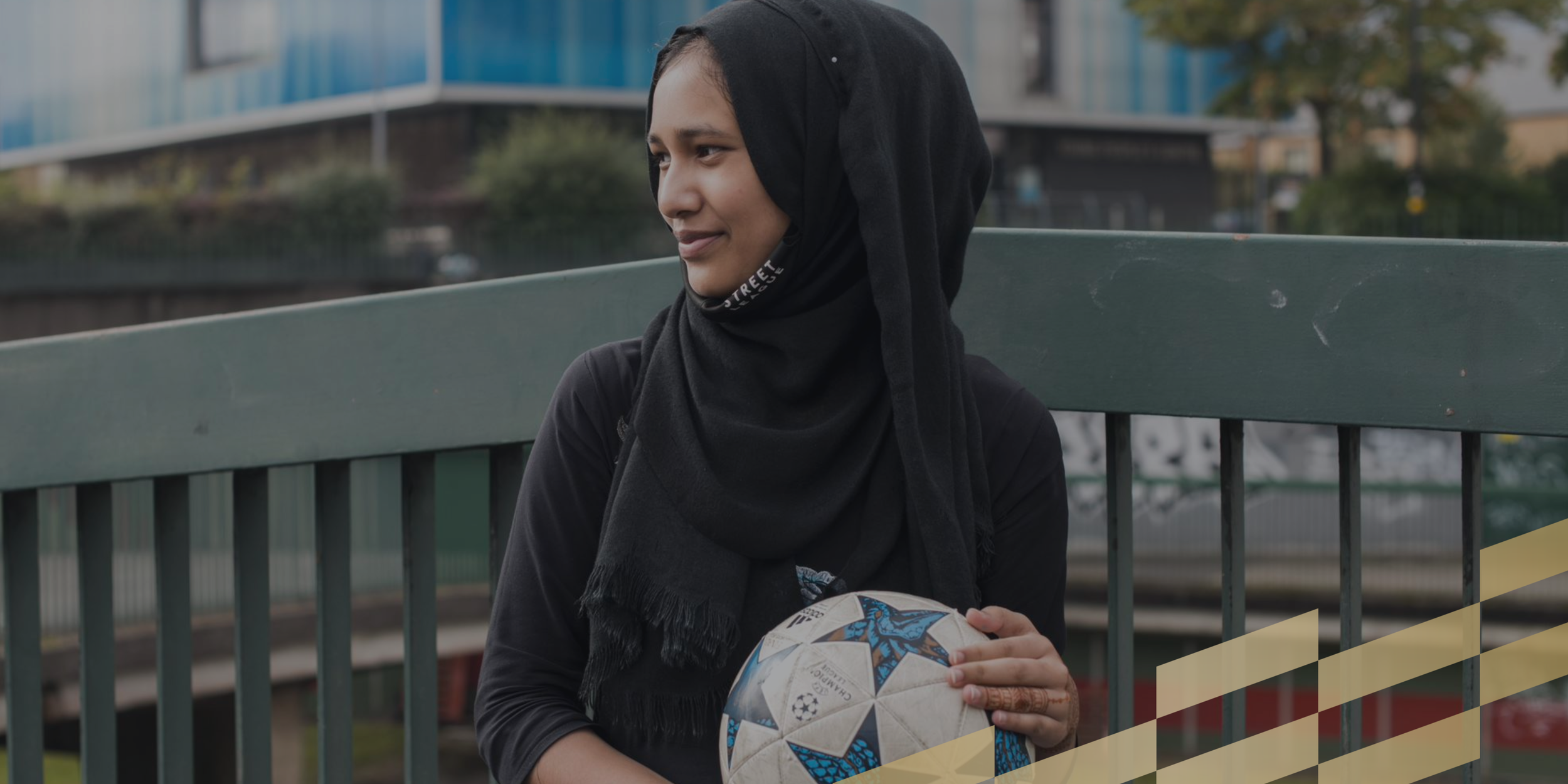 A woman wearing a black hijab and black shirt sitting on a park bench, holding a soccer ball with the UEFA Champions League logo, with a modern building and trees in the background.