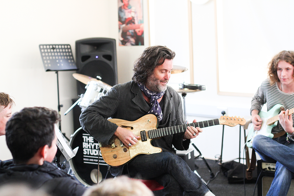 A man with curly hair and a beard playing an electric guitar in a music class, with students and musical equipment around him.