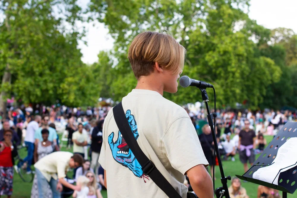 A young person with blonde hair singing or speaking into a microphone at an outdoor event with a large crowd and trees in the background.
