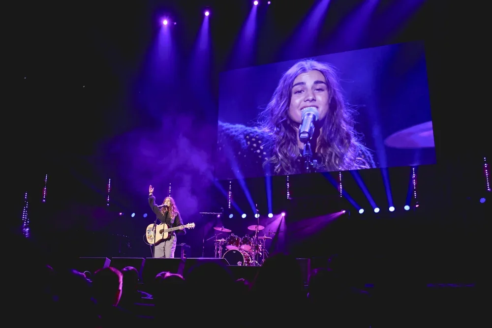 A female singer with long wavy hair performing on stage with a guitar, large screen behind her displaying her close-up, purple stage lighting, and audience in the foreground.
