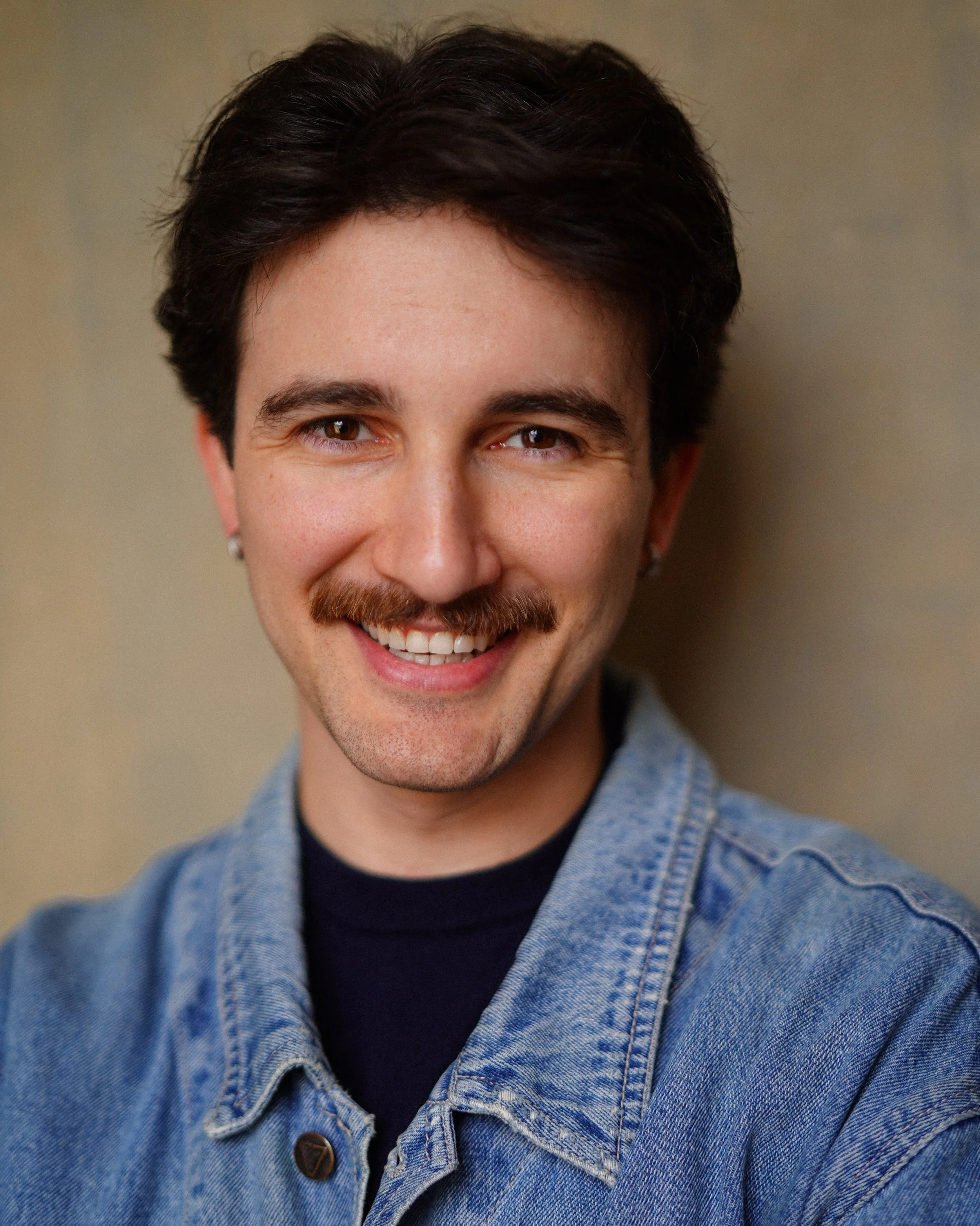 Portrait of a smiling man with dark hair, mustache, wearing a denim jacket and earrings, standing against a neutral background.