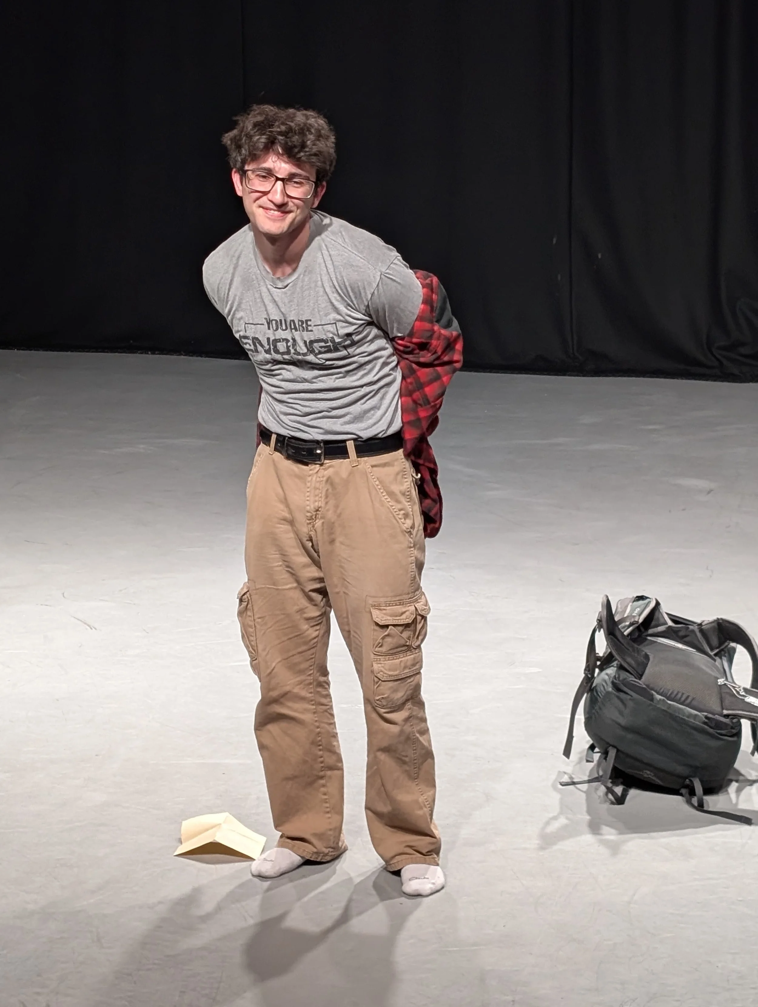 A young man with glasses and messy brown hair stands on a stage, smiling at the camera. He is wearing a gray t-shirt, tan cargo pants, and white socks. There is a black backpack on the stage floor to his right, and a paper airplane is near his feet.
