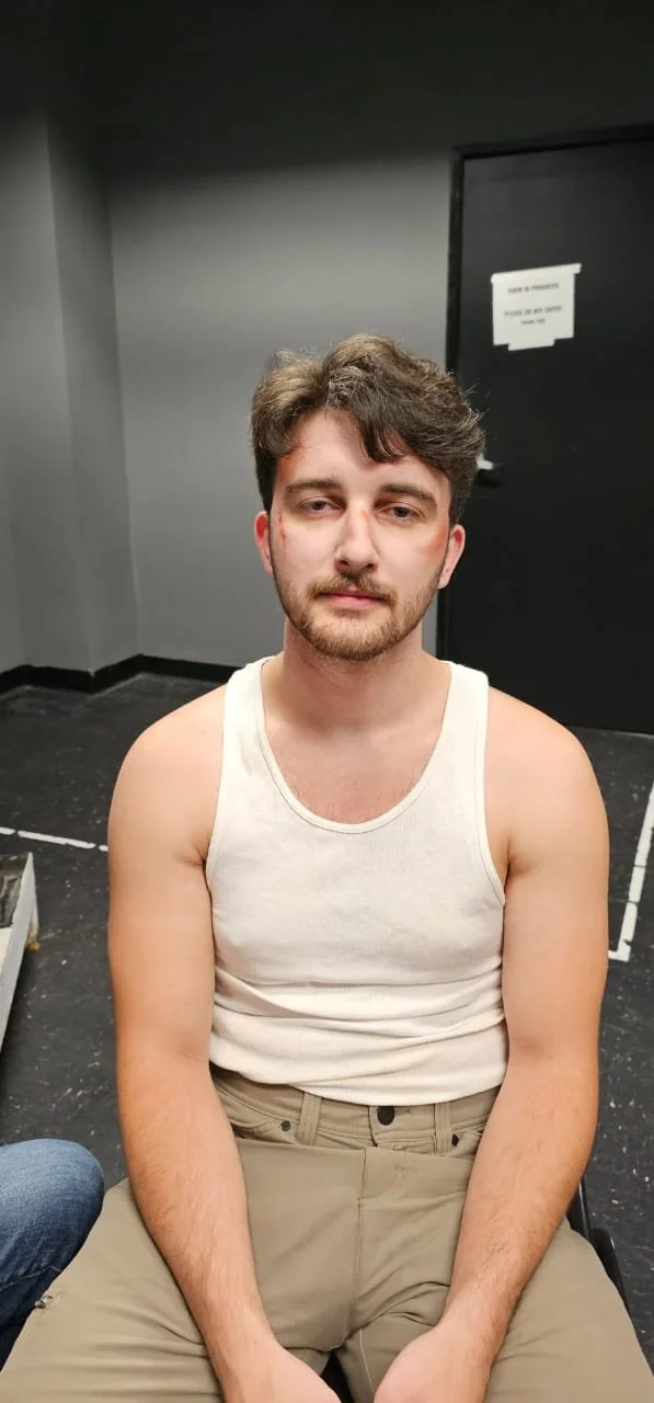 A young man with light skin, brown hair, and a beard, wearing a sleeveless white shirt and beige pants, sitting indoors in front of gray and black walls.