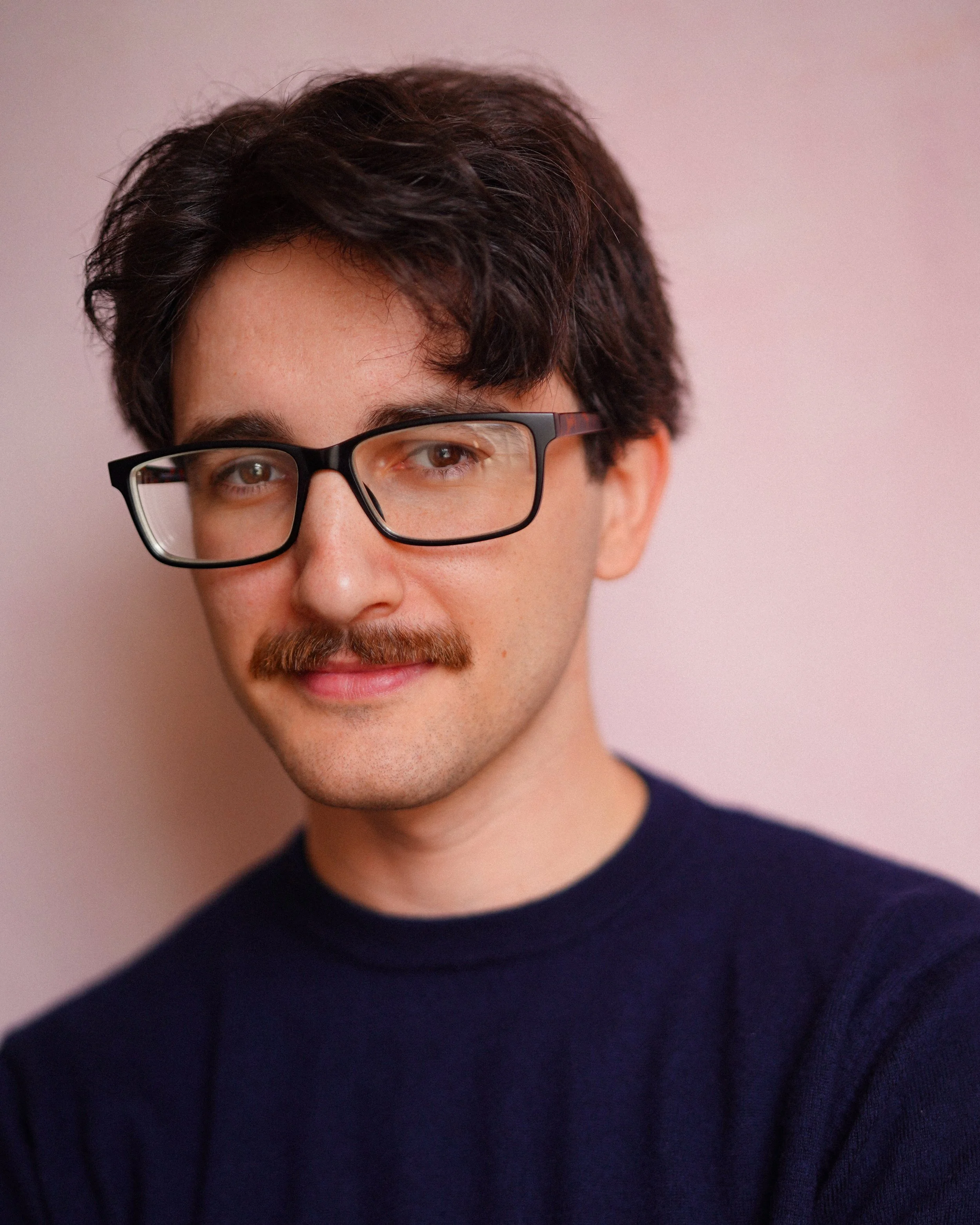 A young man with dark hair, glasses, a mustache, and a beard wearing a dark sweater, standing against a pink background.