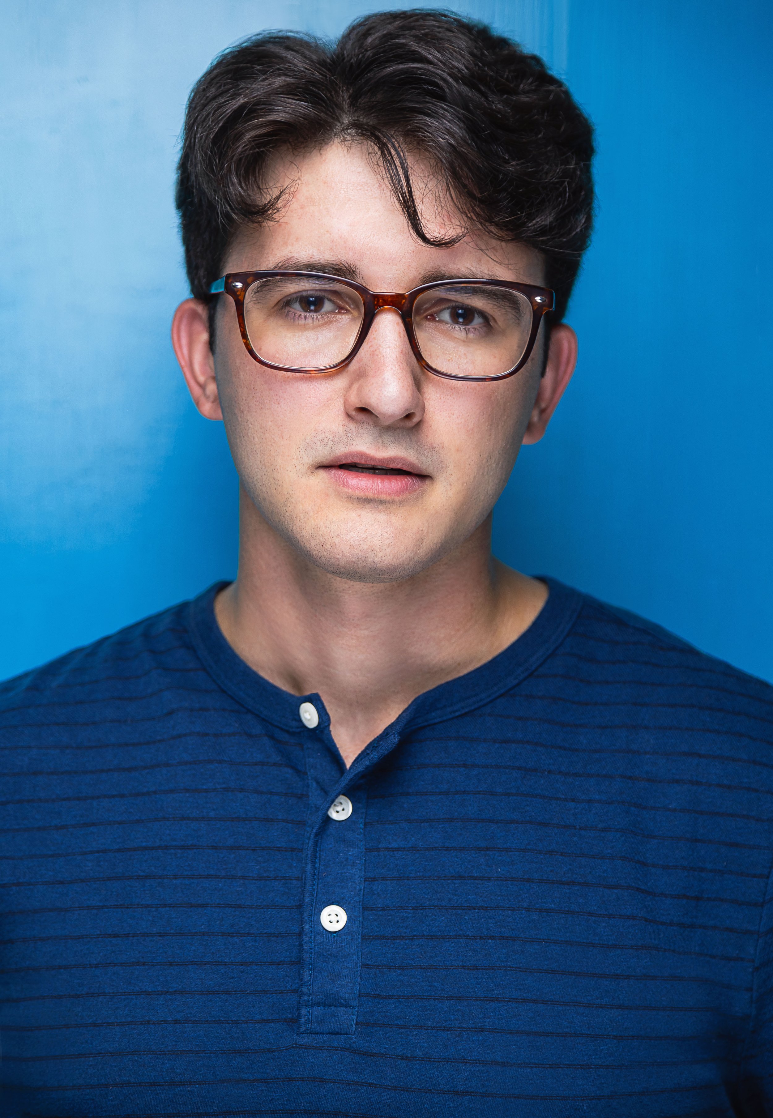 A young man with dark hair, wearing glasses and a blue striped shirt, standing in front of a blue background.