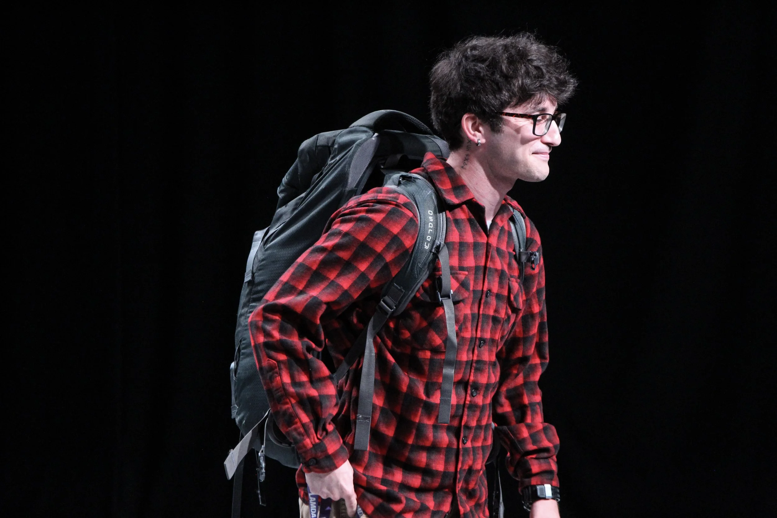 Man with dark curly hair, glasses, and a red and black plaid shirt carrying a large backpack against a black background.