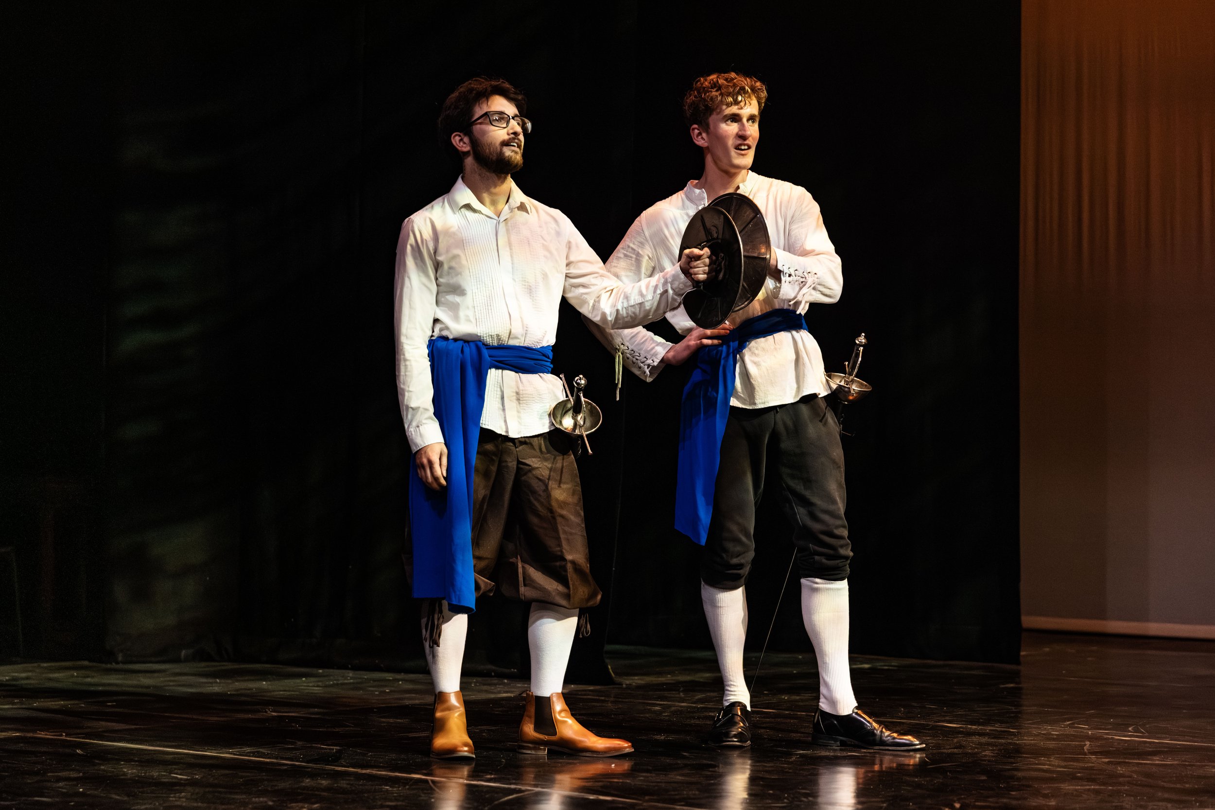 Two men dressed in historical costumes, standing on a stage, holding musical instruments, likely participating in a theatrical performance.