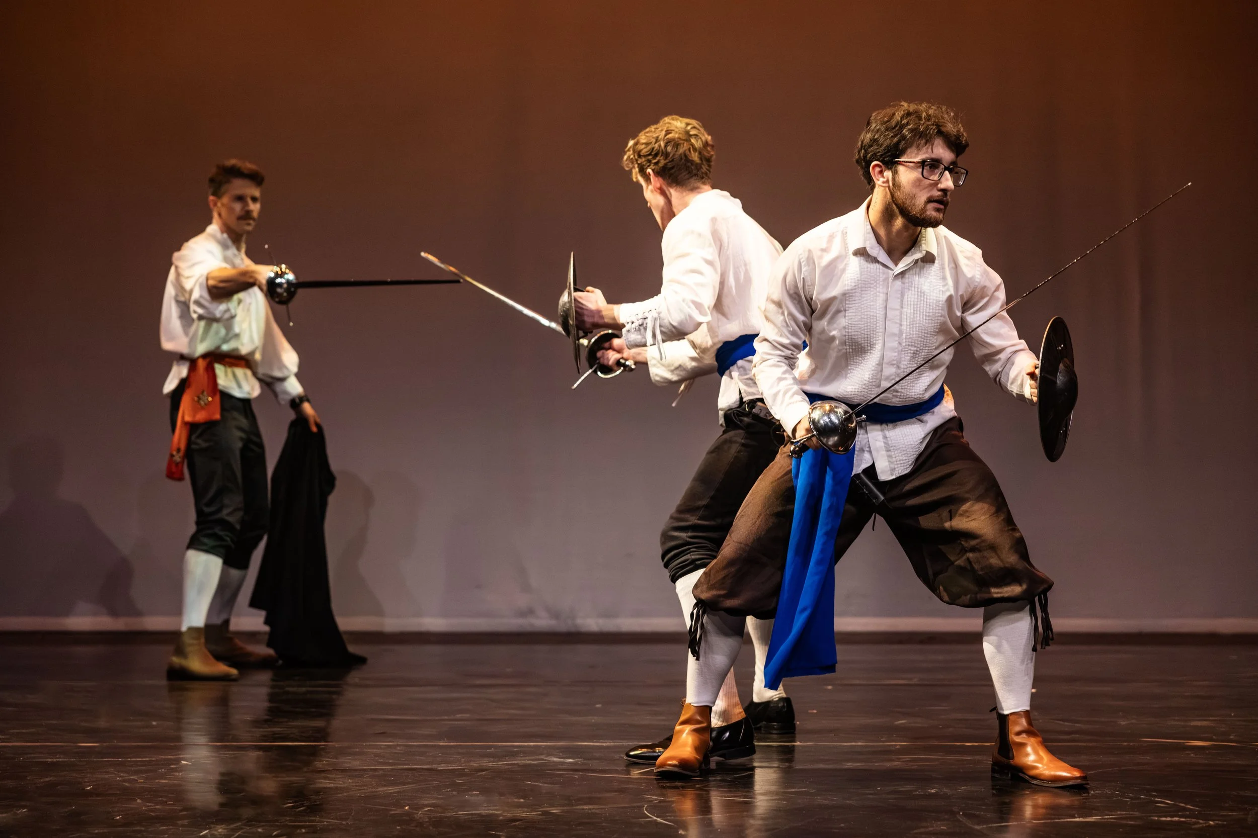 Three men dressed in fencing gear engaged in a fencing match on stage.
