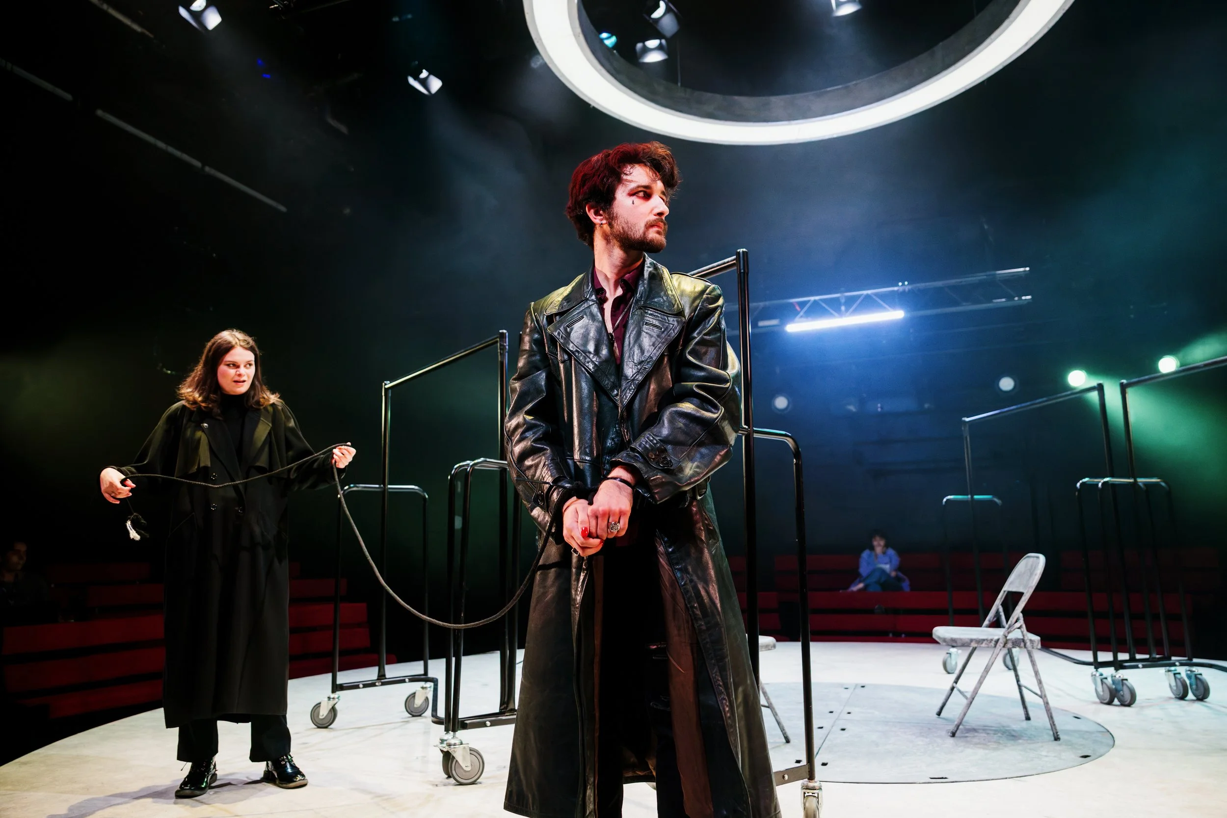 Theatrical scene with two actors on stage; one man in a leather trench coat standing at center, a woman in dark clothing holding a cord behind him, with a person sitting in the background.