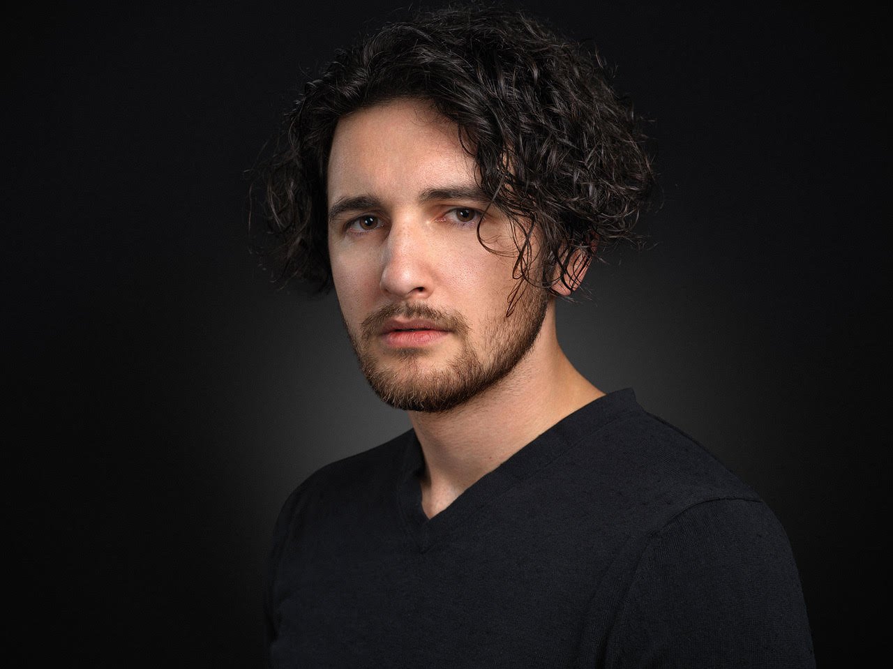 Portrait of a young man with wavy dark hair, beard, wearing a black shirt, against a dark background.