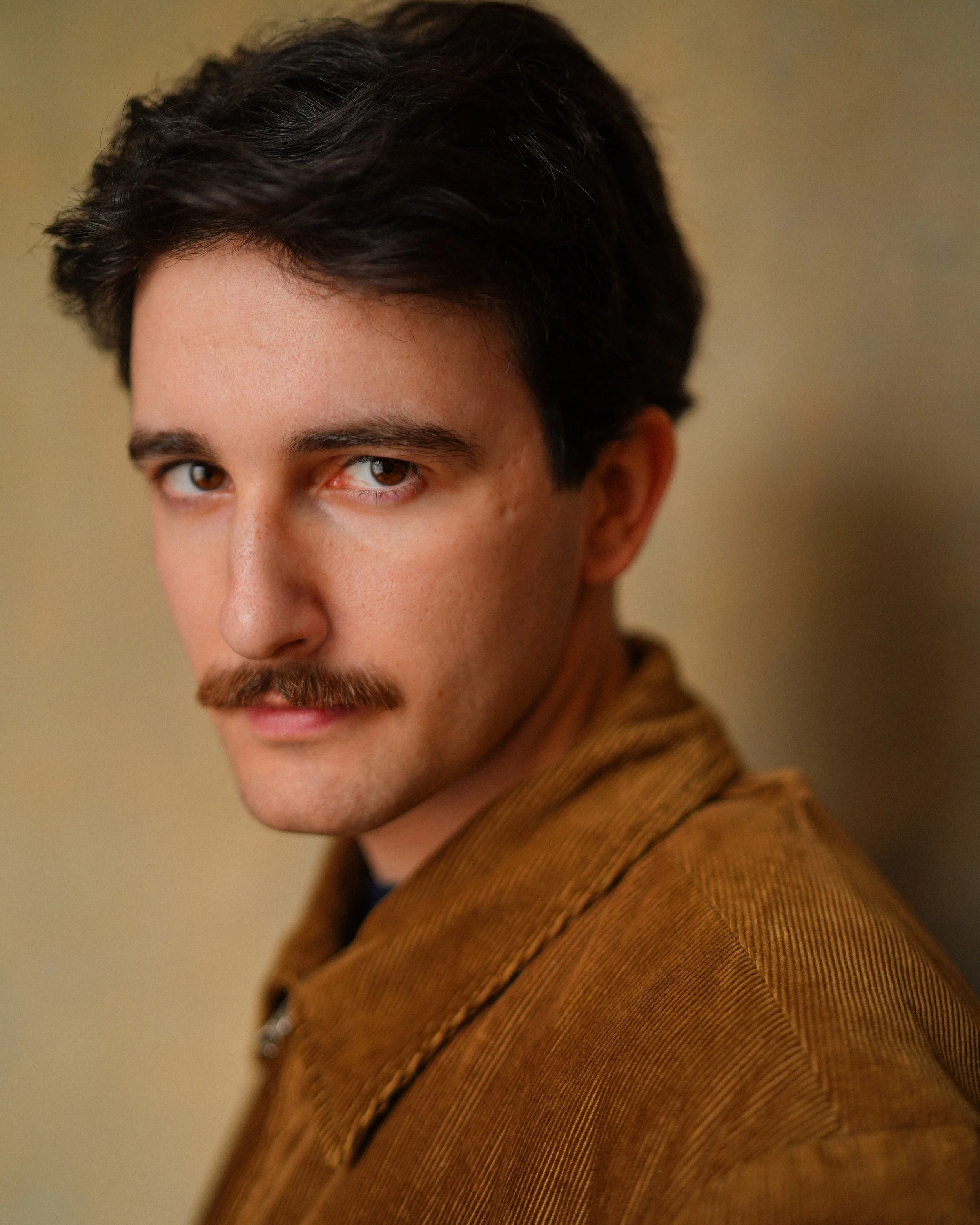 A young man with dark hair, a mustache, light skin, and wearing a brown jacket, looking at the camera with a neutral expression.
